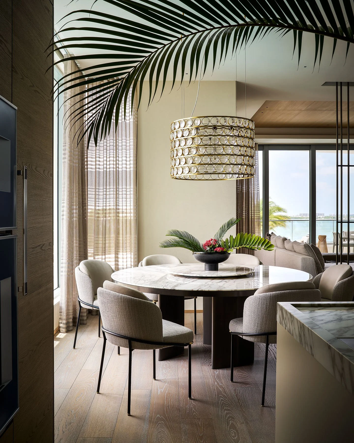 Luxury dining room with round marble table, sculptural chandelier, and waterfront balcony views.