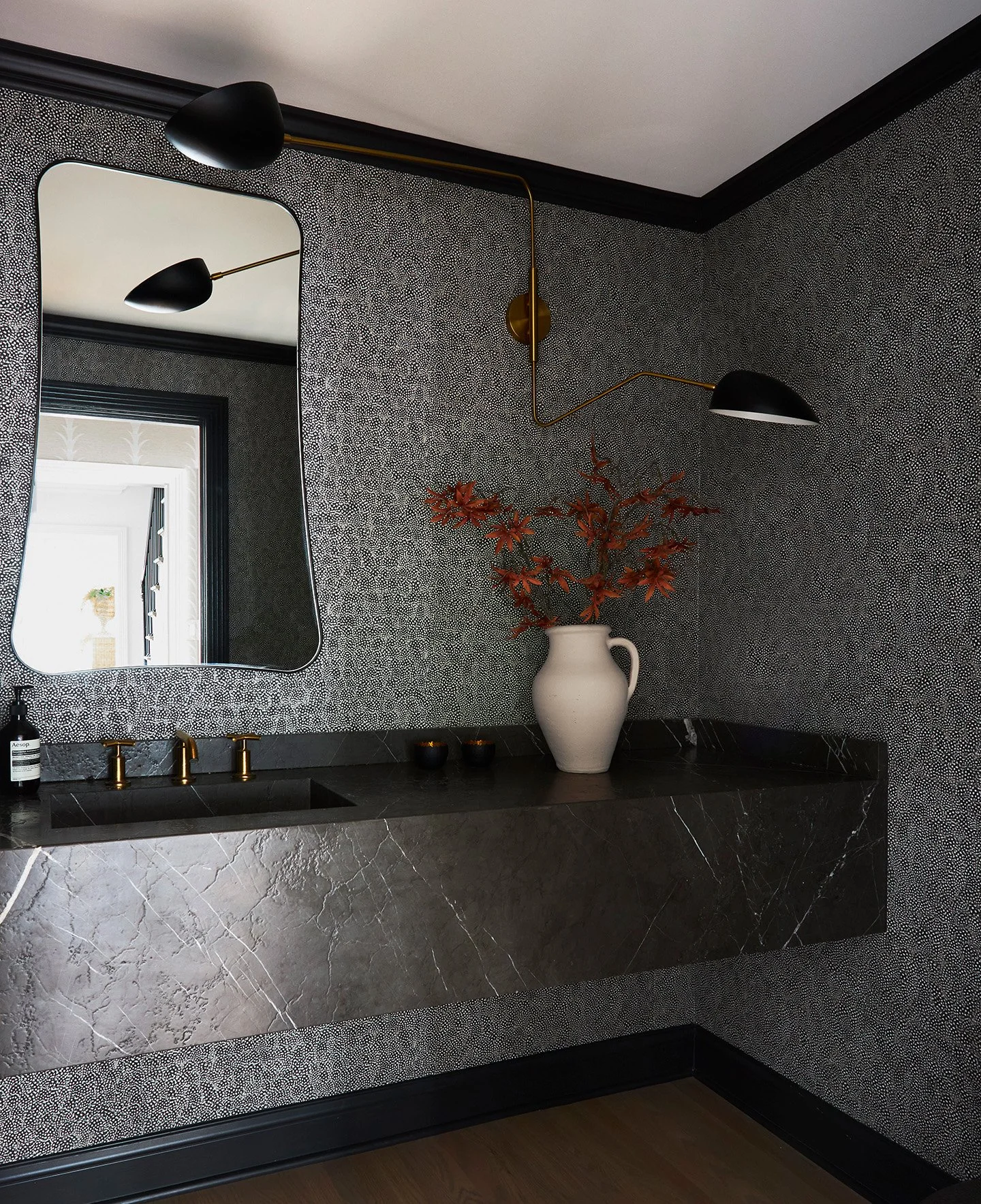 Moody powder room with black marble floating sink, brass fixtures, sculptural mirror, and patterned walls.