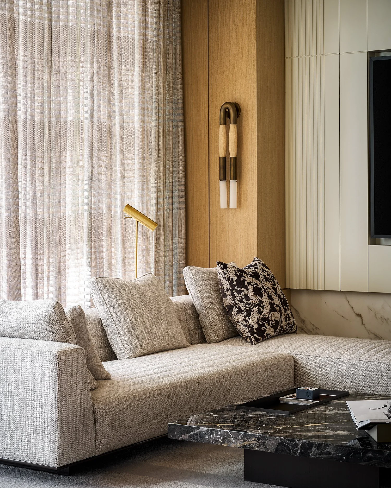 Modern living room with beige sectional sofa, various pillows, a book on a black marble coffee table, floor-to-ceiling curtains, a floor lamp, decorative wall art, and built-in wall cabinetry.