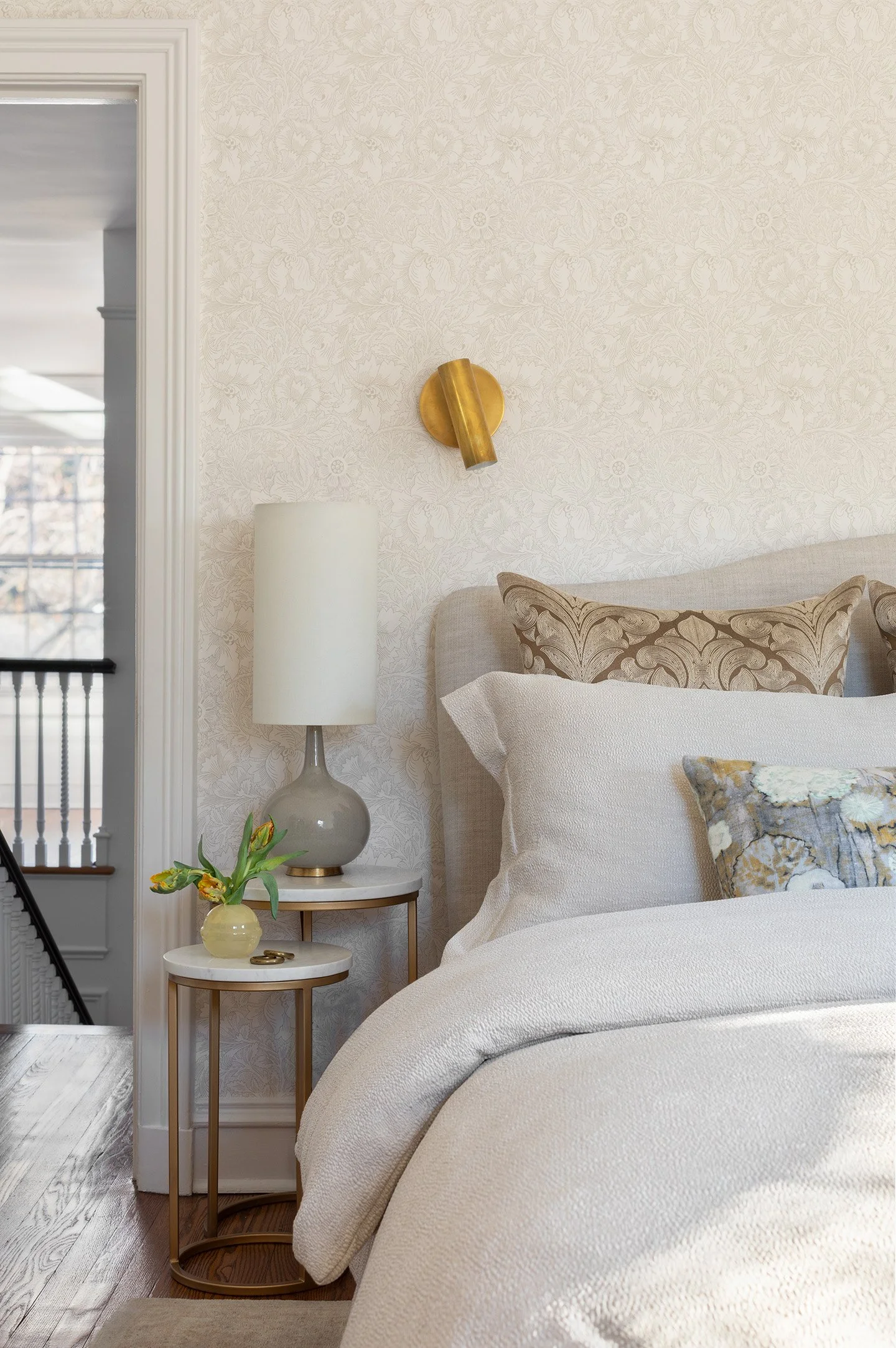 Neutral bedroom with textured wallpaper, brass wall sconce, layered bedding, and round marble side tables.