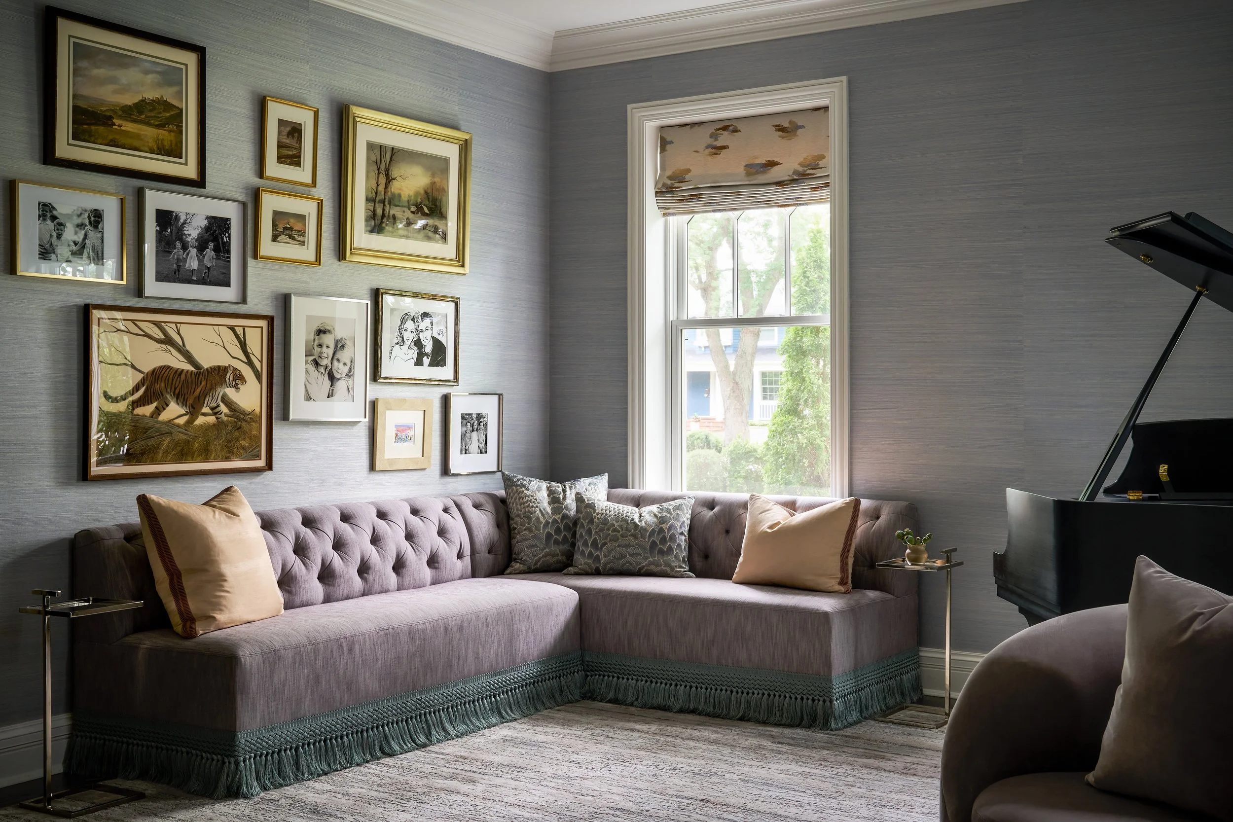 A tailored corner banquette with tufted upholstery and fringe detail, set against a curated gallery wall of framed artwork and family photos, with a grand piano adding elegance to the space.