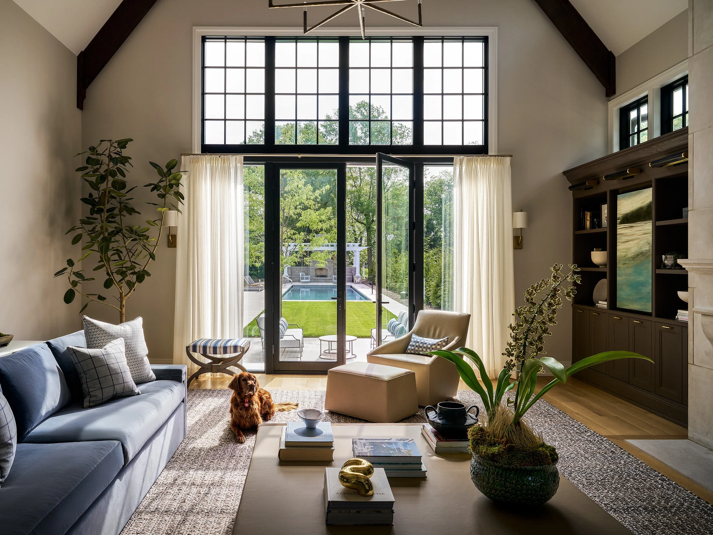 Bright living room with large black-framed windows, soft neutral seating, indoor plants, and a view of a landscaped backyard pool.