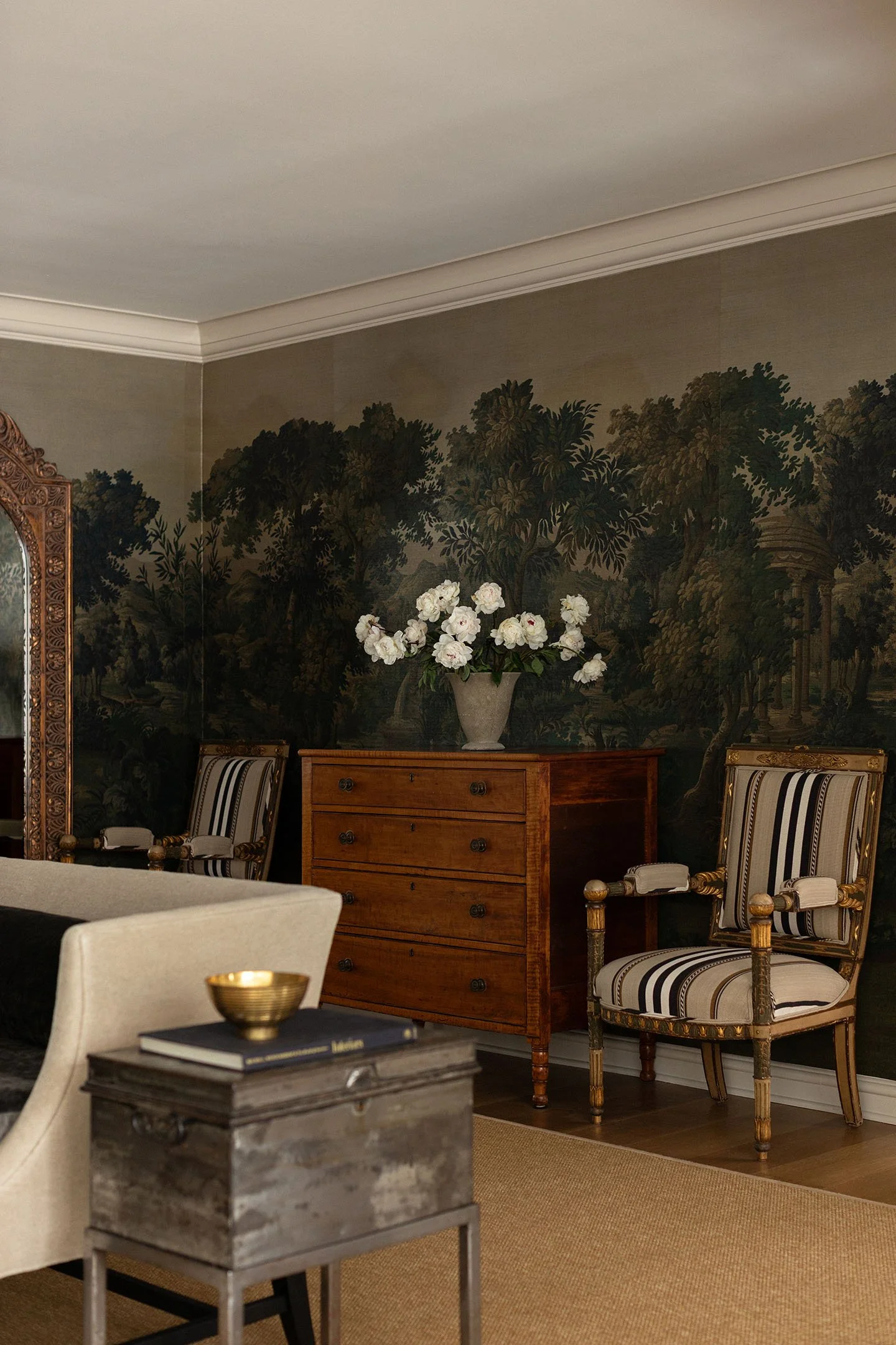 Antique wood chest with striped armchairs set against dramatic scenic mural wallpaper in a classic interior.