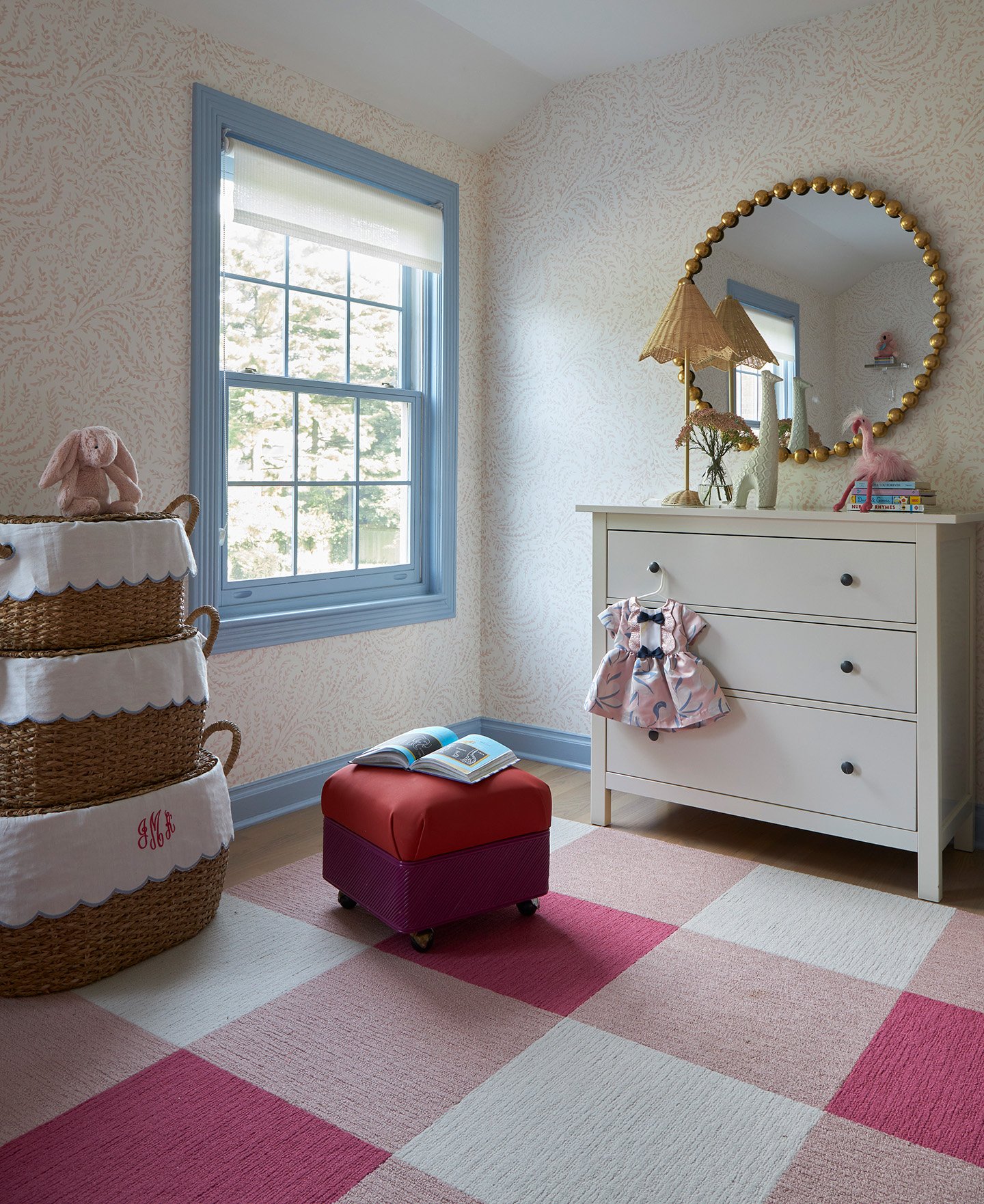 Girls bedroom with blue window trim, white dresser, round gold mirror, and pink checkered rug.