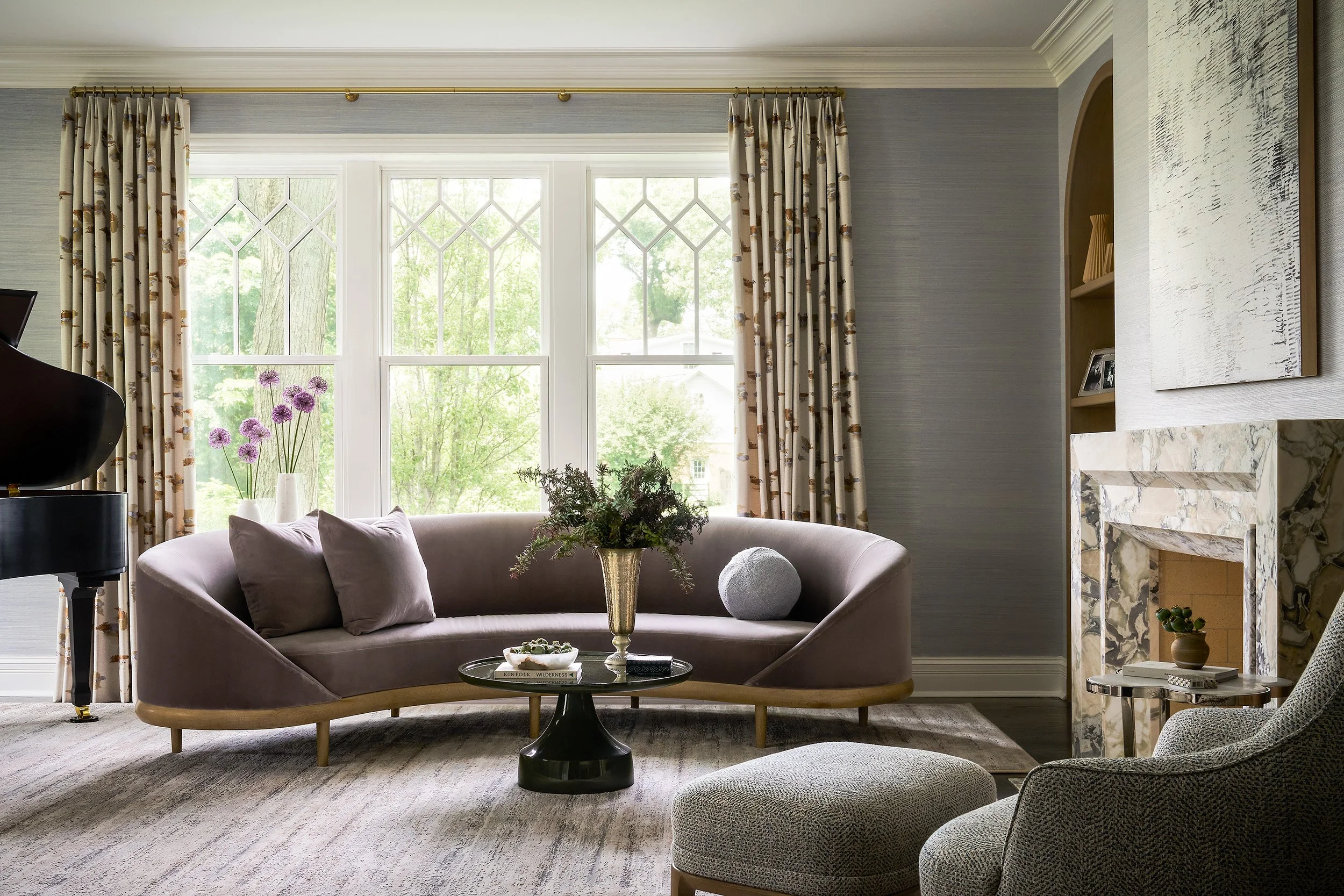A softly lit living space with a curved blush sofa centered beneath tall windows dressed in patterned drapery, complemented by a glass coffee table and layered neutral textures.