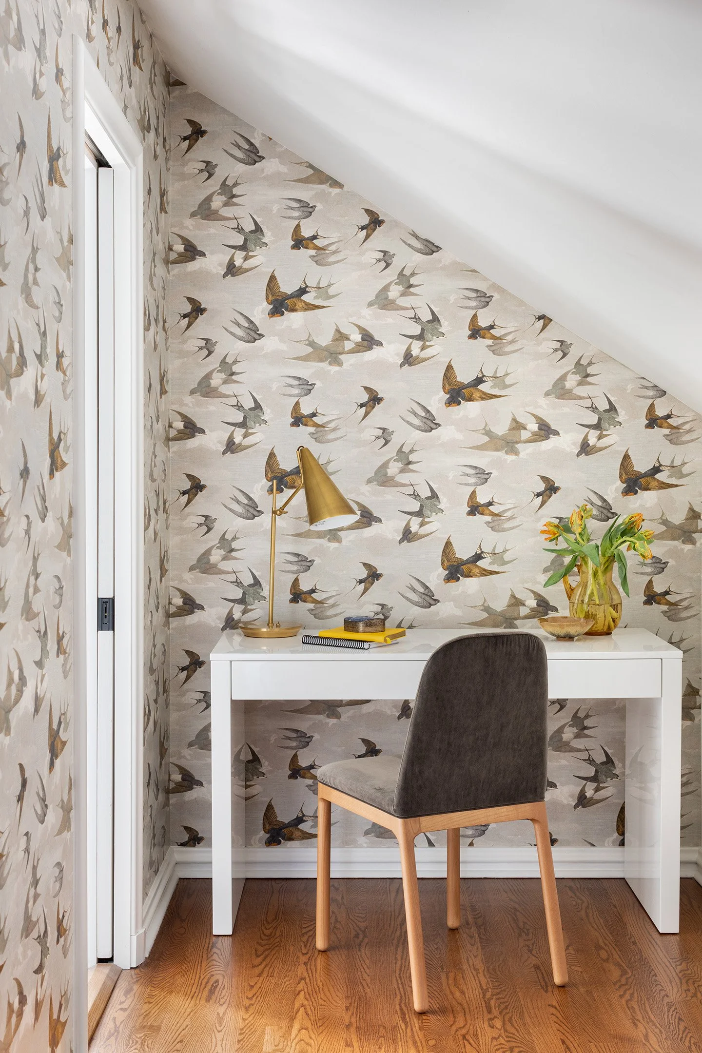 Home office nook with bird patterned wallpaper, white desk, brass lamp, and upholstered chair.