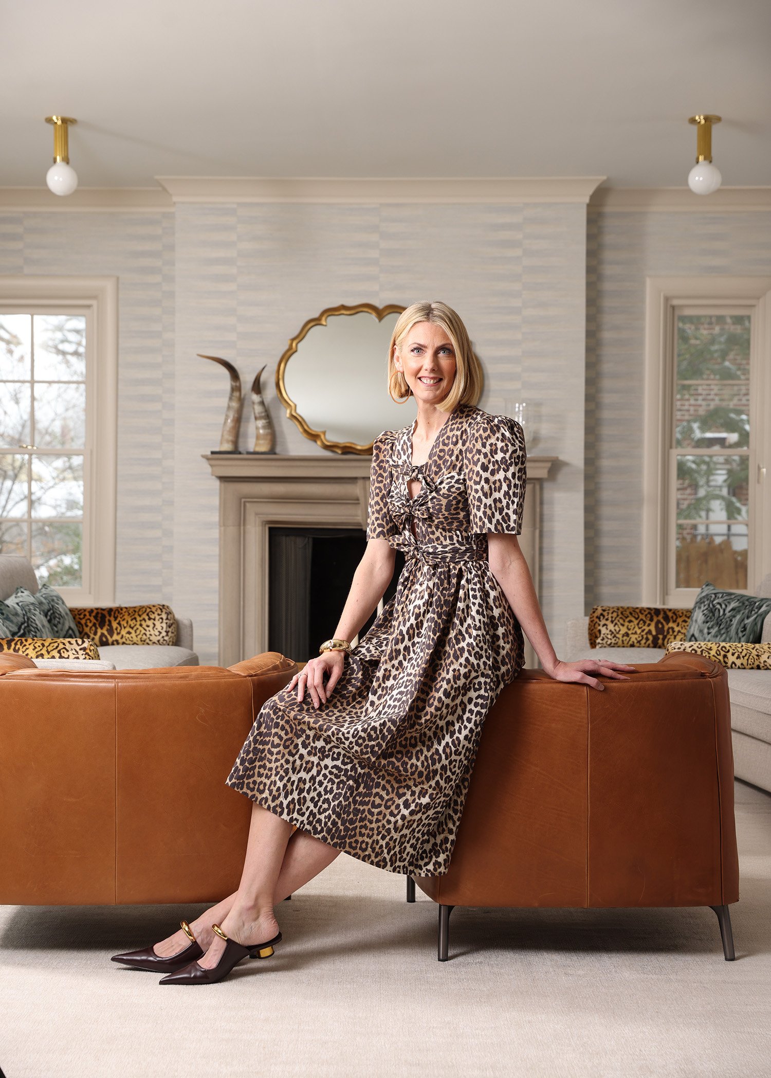 A woman with blonde hair wearing a leopard print dress, sitting on a brown leather couch in a decorated living room with a fireplace, mirror, and windows.