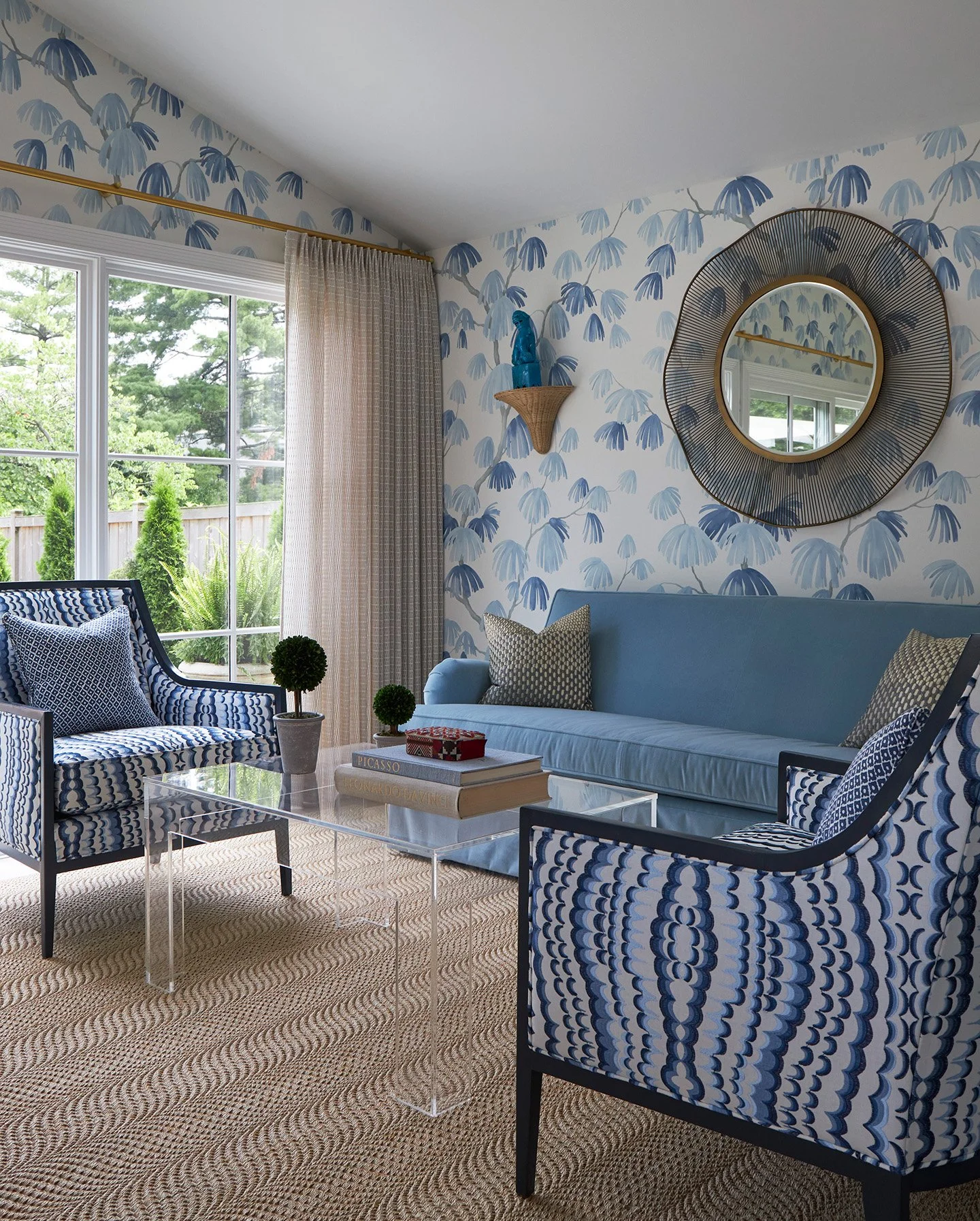 Living room with blue botanical wallpaper, matching sofa and chairs, round mirror, and acrylic coffee table.