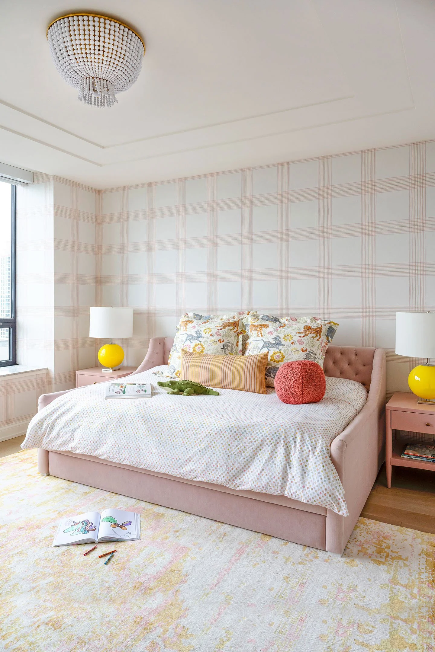 Girls bedroom with blush upholstered bed, plaid wallpaper, colorful bedding, yellow bedside lamps, and soft pastel area rug.