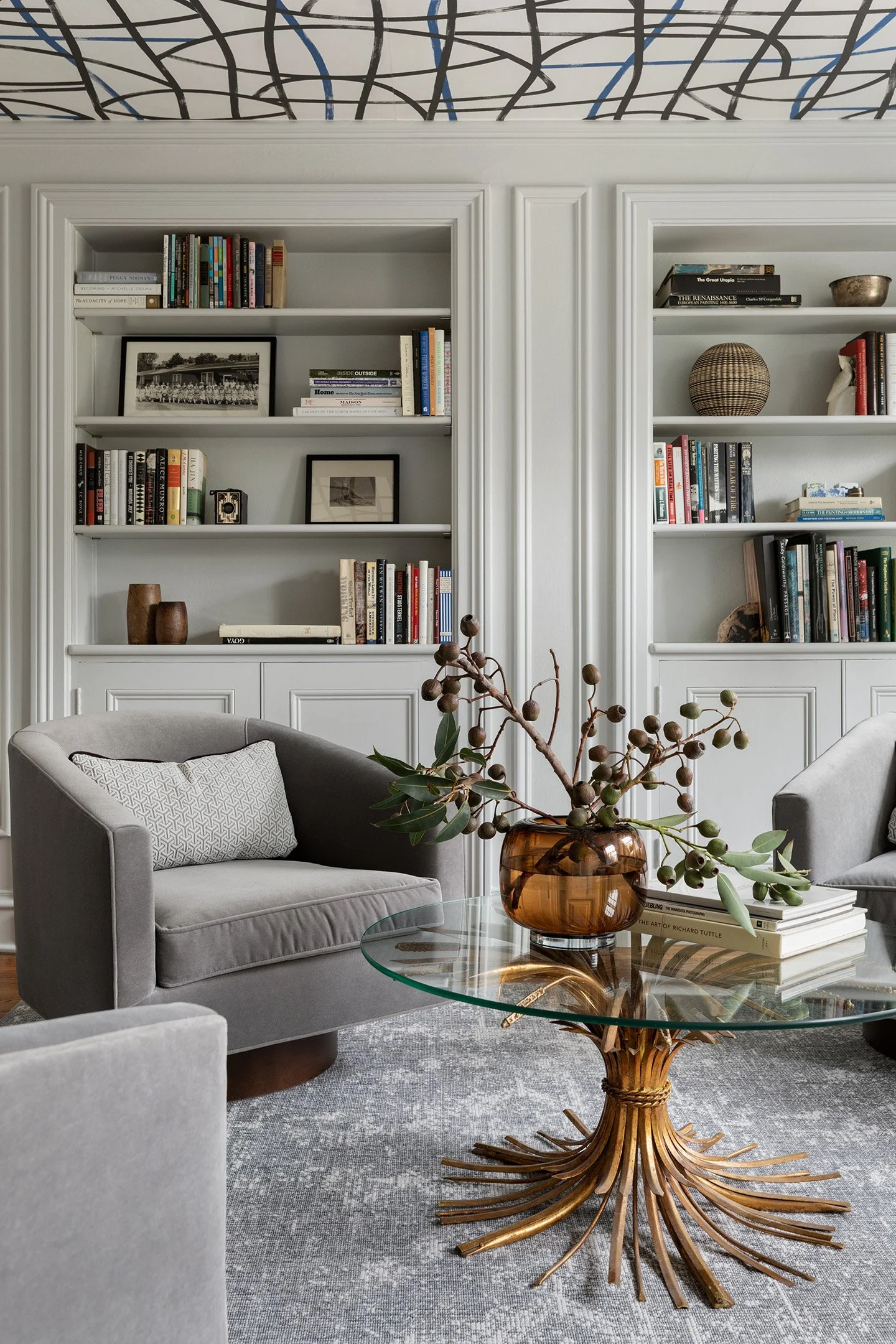 Custom built-in bookshelves behind gray lounge chairs and sculptural glass coffee table.