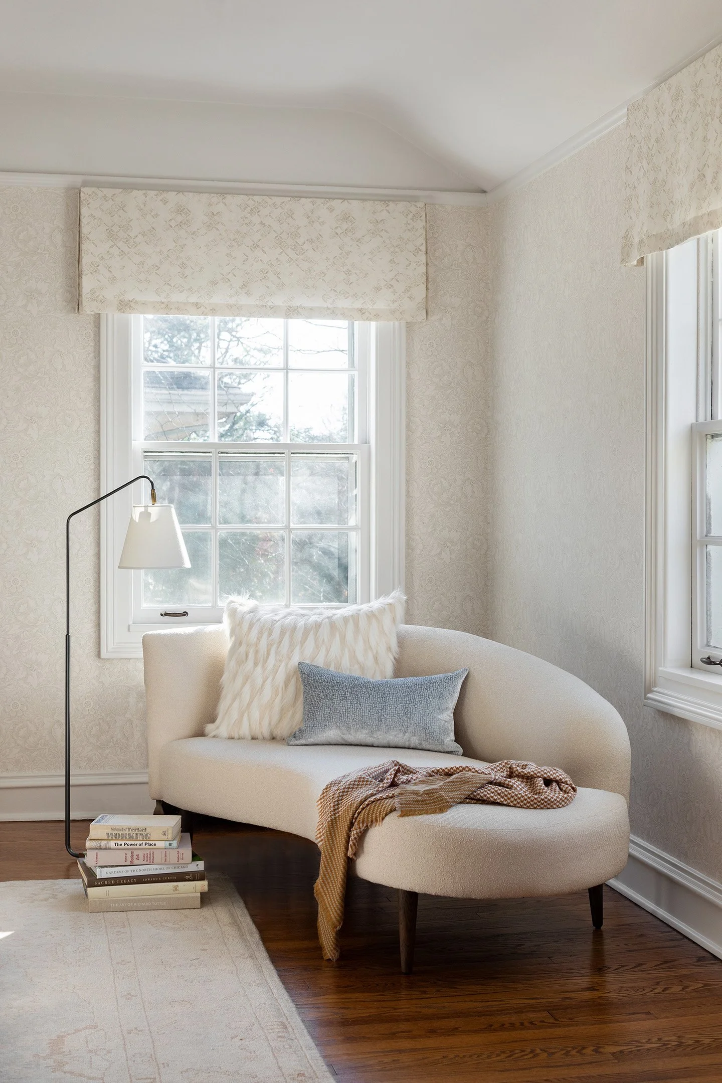Cozy reading nook with curved cream settee, floor lamp, patterned wallpaper, and soft natural light.