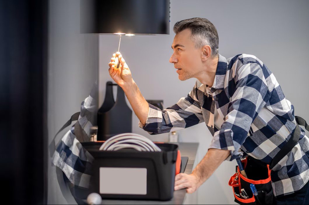 A man wearing a plaid shirt and a tool belt inspects electrical wiring with a flashlight in an indoor setting.