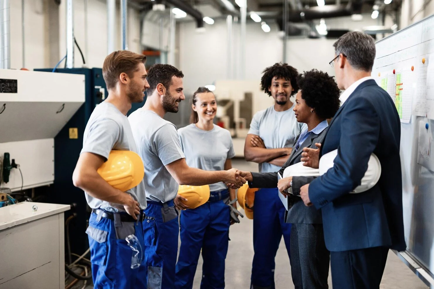 A group of workers and managers in a factory, with one worker shaking hands with a manager. The workers are holding yellow safety helmets, and the background shows industrial equipment and charts on a whiteboard.