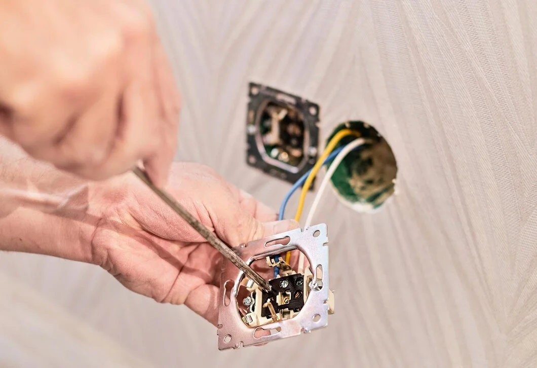 A person is installing or repairing an electrical switch outlet in a wall with a screwdriver, with electrical wires visible in a hole in the wall.