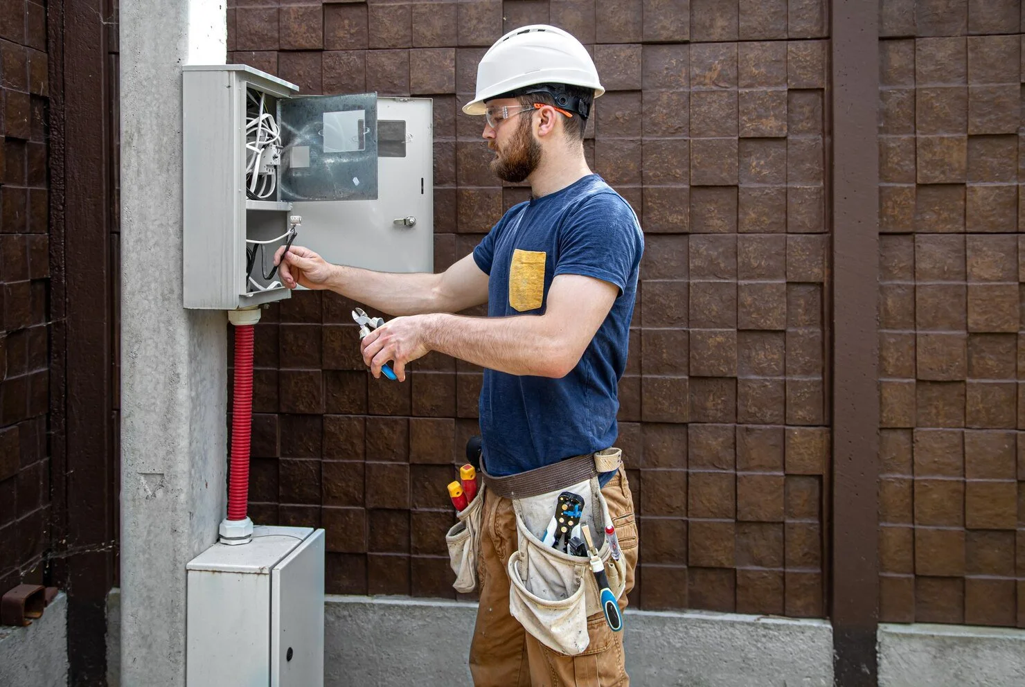 A male electrician wearing a cowboy hat, safety goggles, and a tool belt working on an electrical panel outdoors against a brick wall.