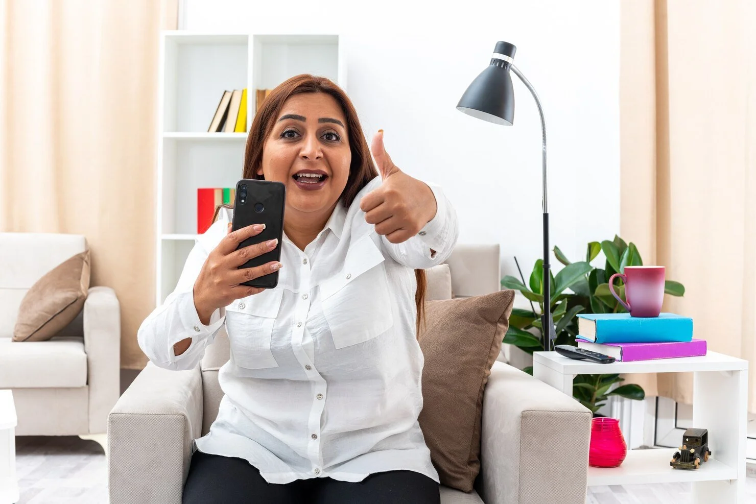 A woman sitting on a beige sofa holding a smartphone and giving a thumbs up in a bright living room with books, a pink mug, books on a table, a lamp, and houseplants.