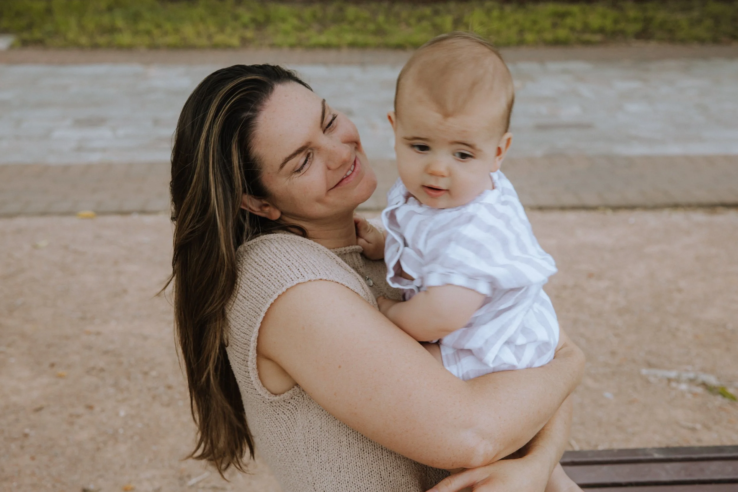 A woman holding a young child outside near a beach with sand and water, smiling and looking at the child.