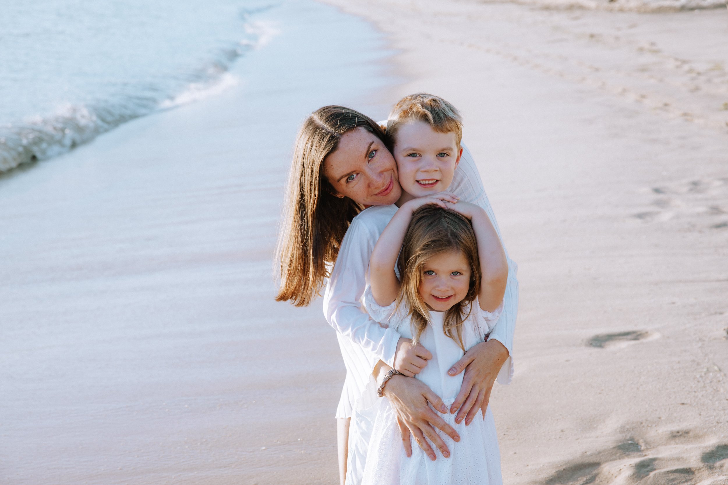 A woman with long brown hair and two young girls, one with long blond hair and one with shoulder-length light brown hair, standing on a sandy beach near the ocean, smiling and embracing each other.