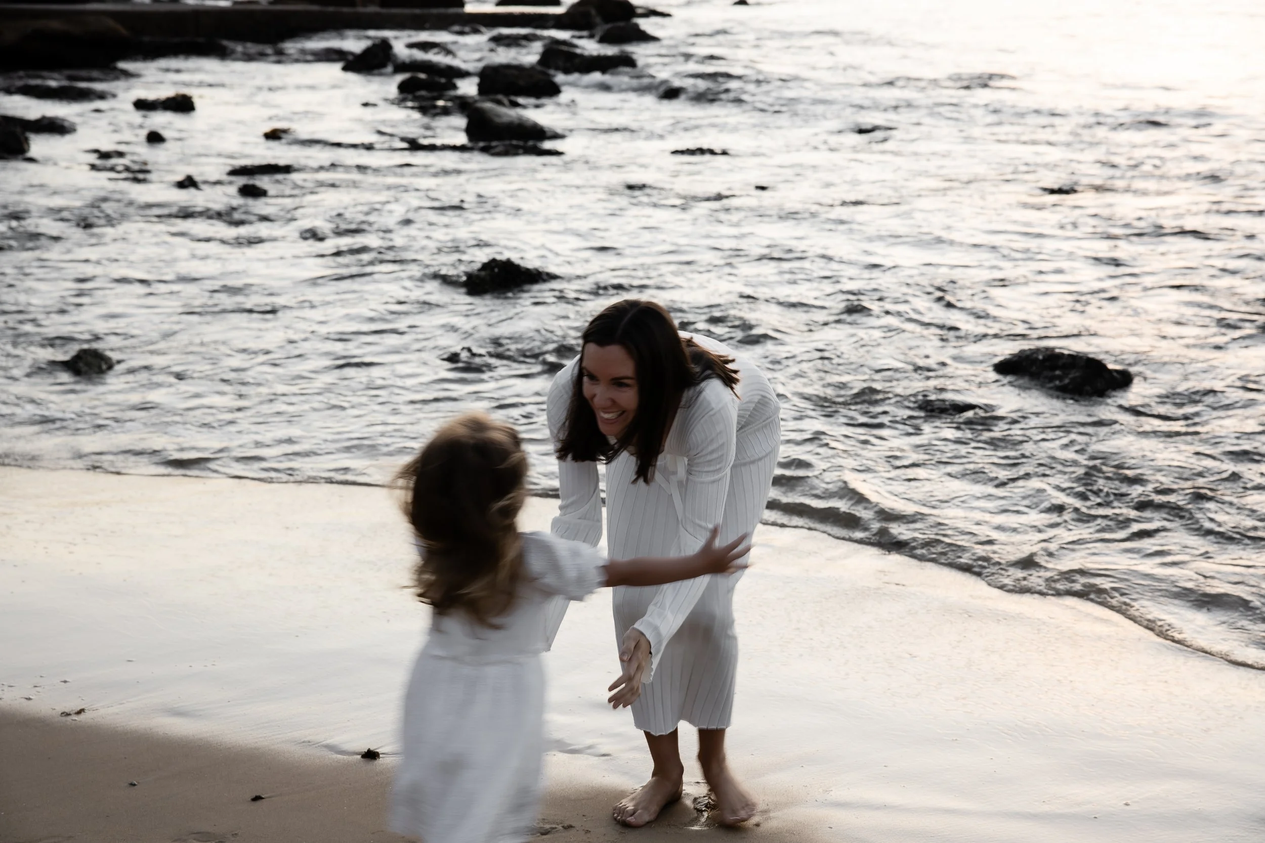 A woman and a young girl, both dressed in white, smiling and holding hands on the beach near the water at sunset.