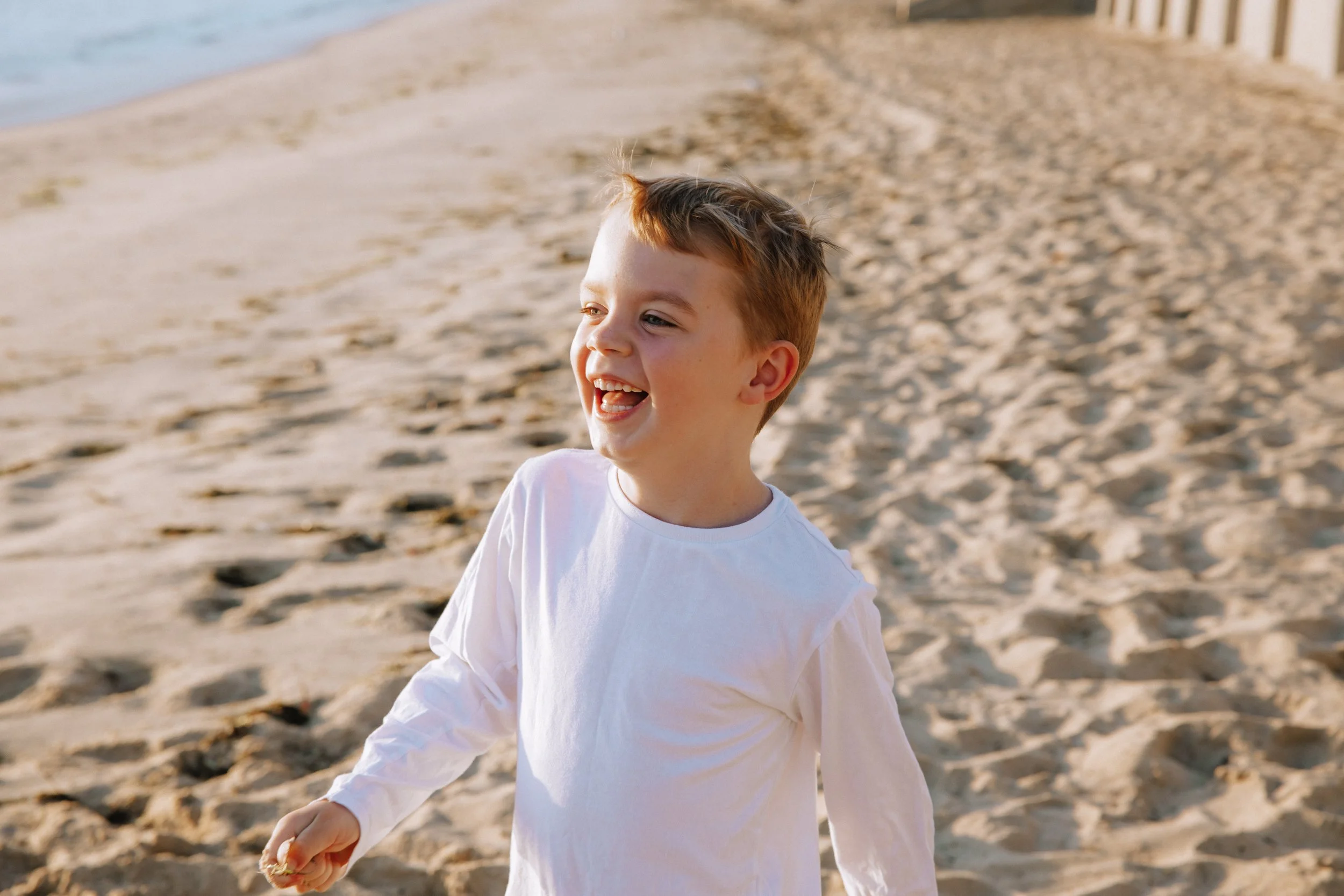 A young boy with short, light brown hair, wearing a white long-sleeve shirt, walking on a sandy beach and smiling.