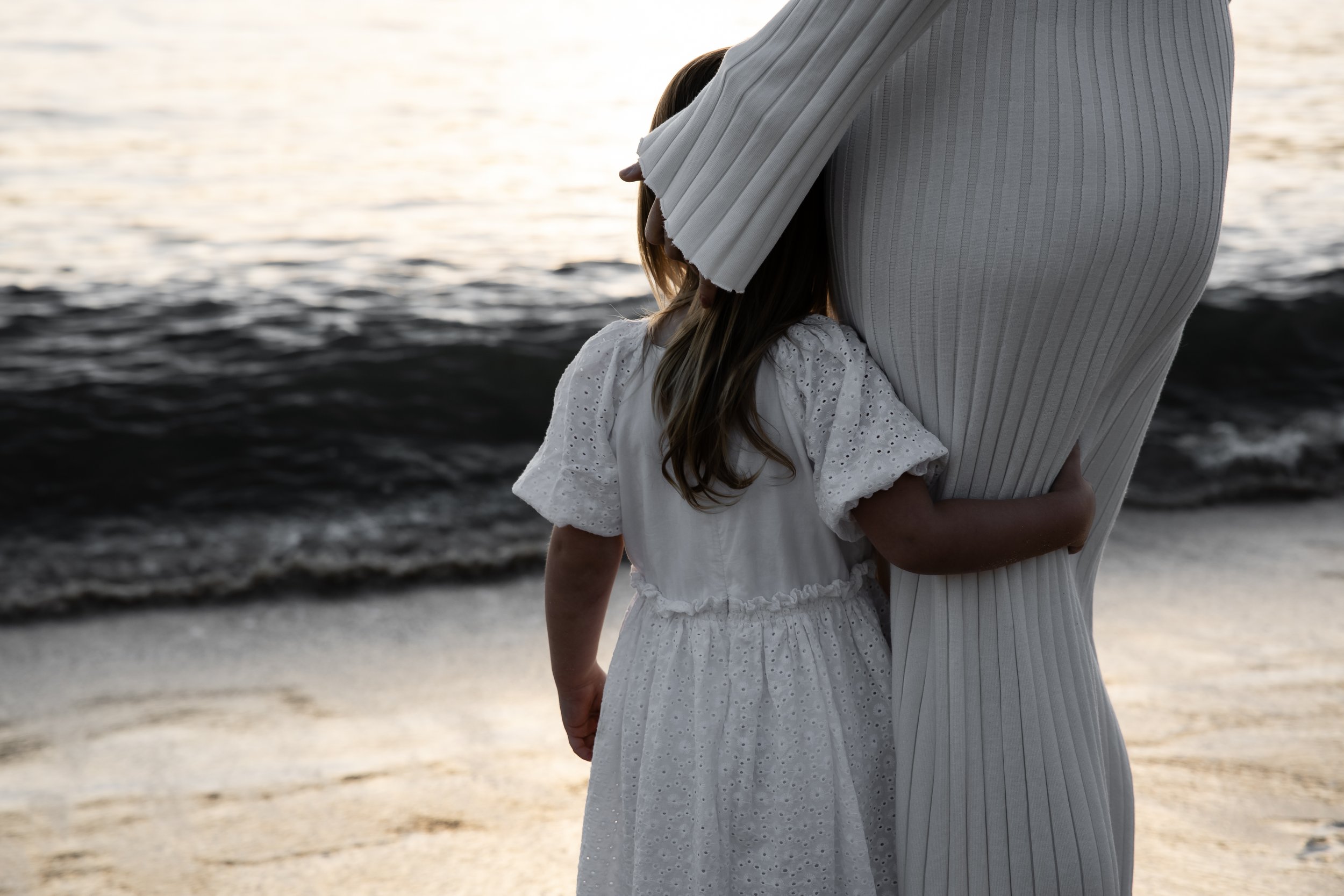 A mother and daughter standing by the ocean at sunset, embracing each other.