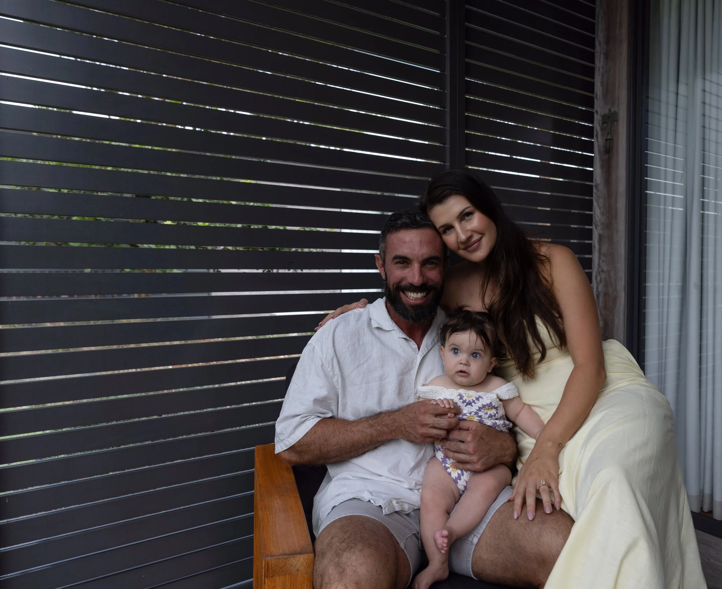 A happy family of three sitting closely together outdoors in front of a dark striped fence. The man has a beard and is wearing a white shirt. The woman has long dark hair and is wearing a light-colored dress. The baby, sitting on the man's lap, looks curious and has dark hair and bright eyes.