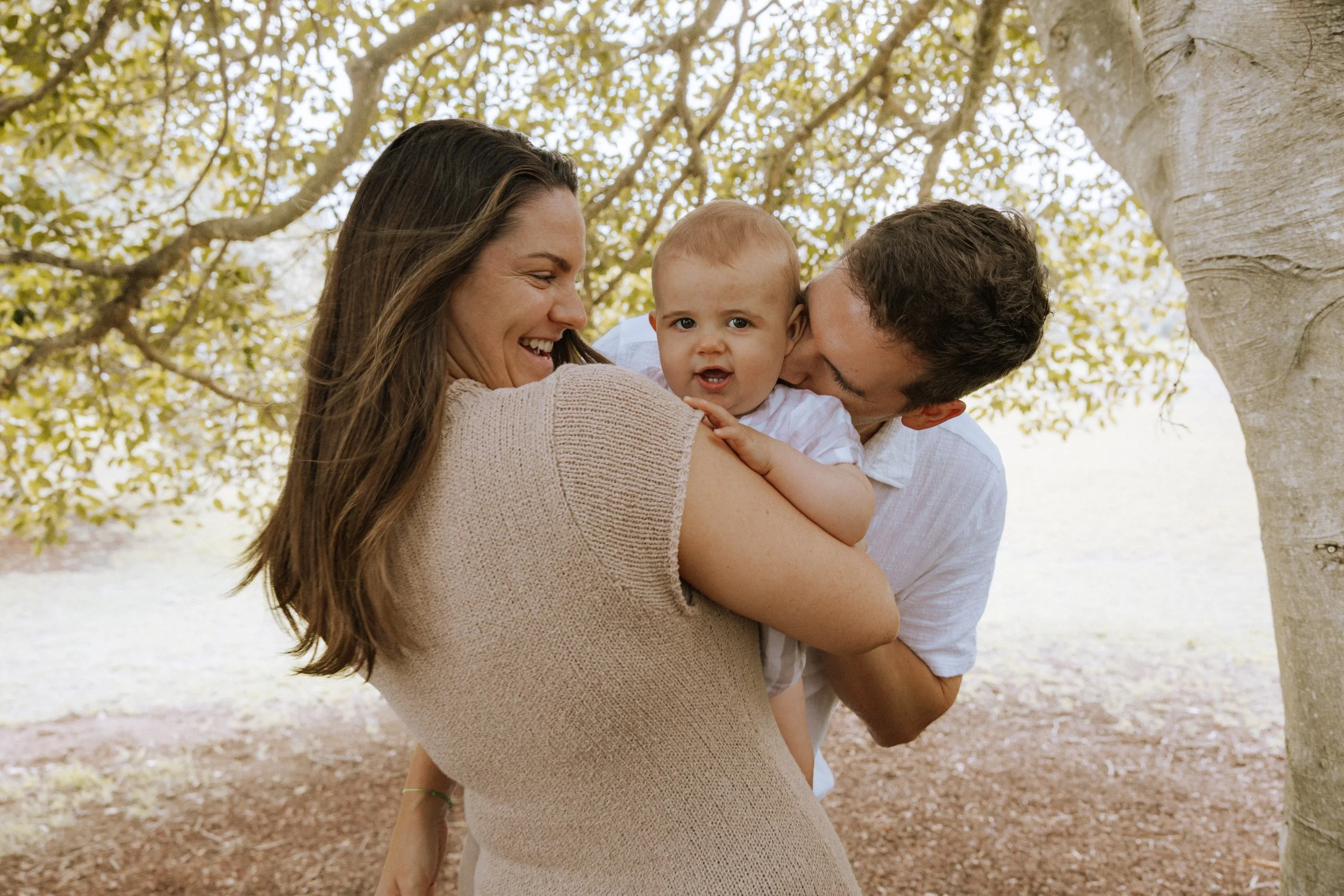 A woman holding a young child outdoors, with a man kissing the child's cheek. All are smiling and under a tree with leaves.