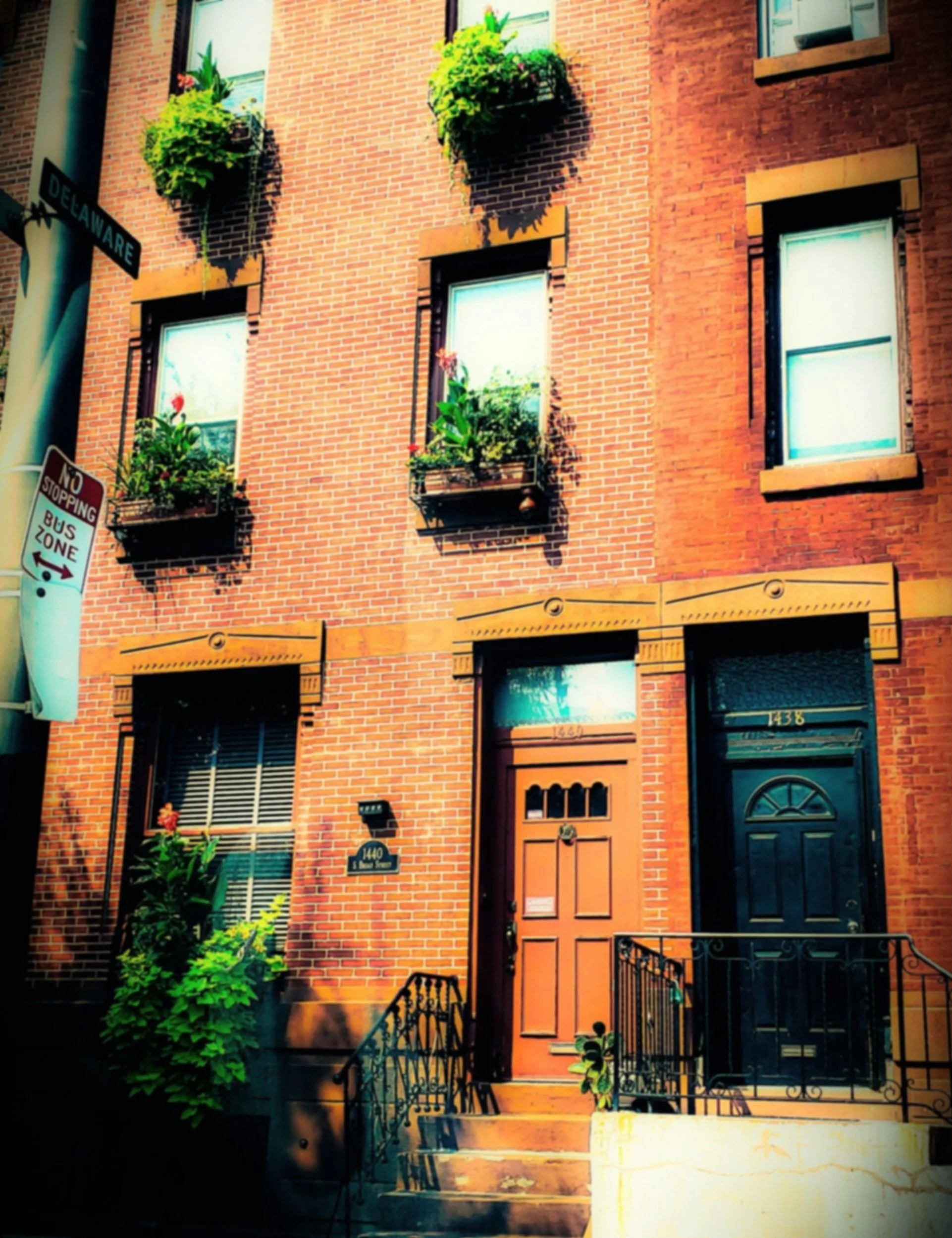 A brick building with six windows and two front doors. Each window has flower boxes with green plants and flowers. The building has a fire escape and stairs leading to the doors. Signs indicate a corner of Delaware Street and a no stopping bus zone.