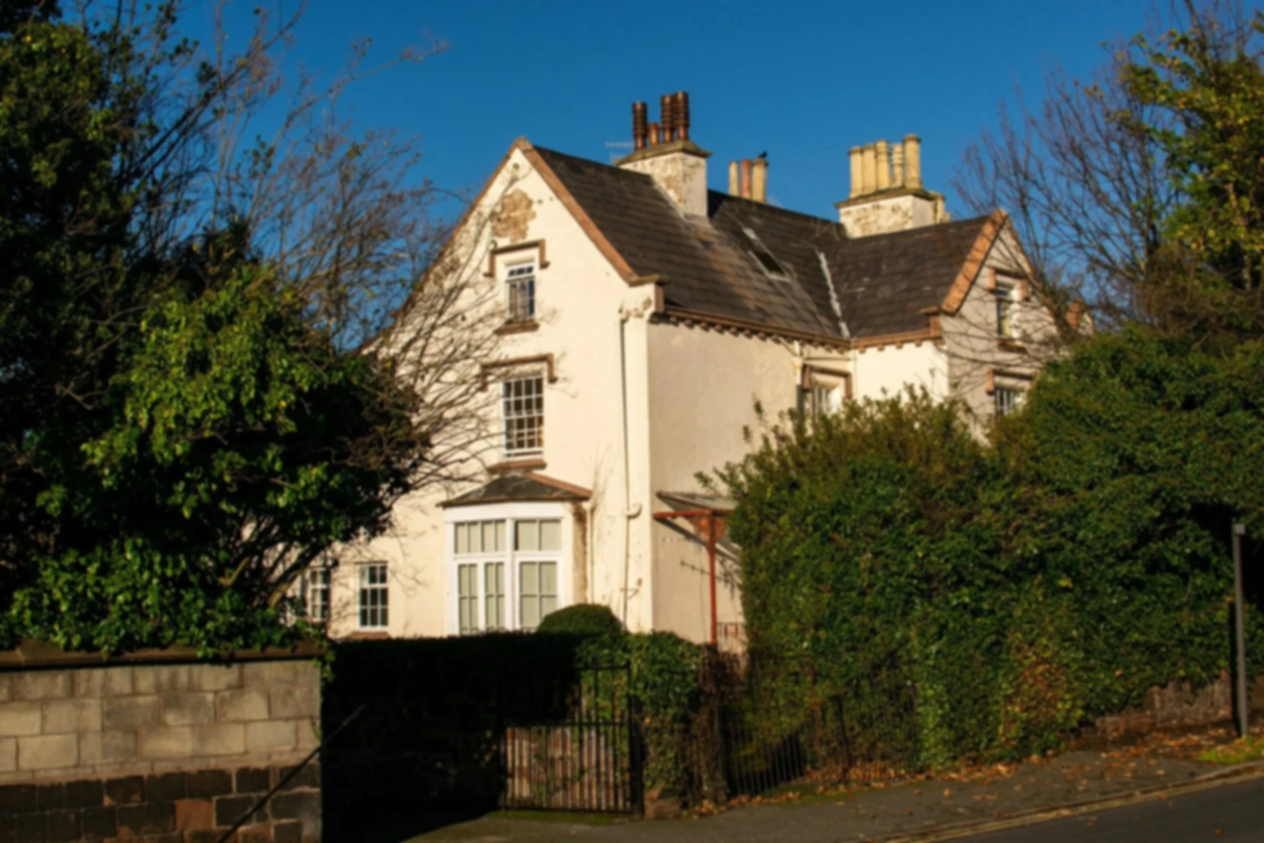 A large, old white house with a steep roof, surrounded by trees and bushes, and a black metal fence in the front.
