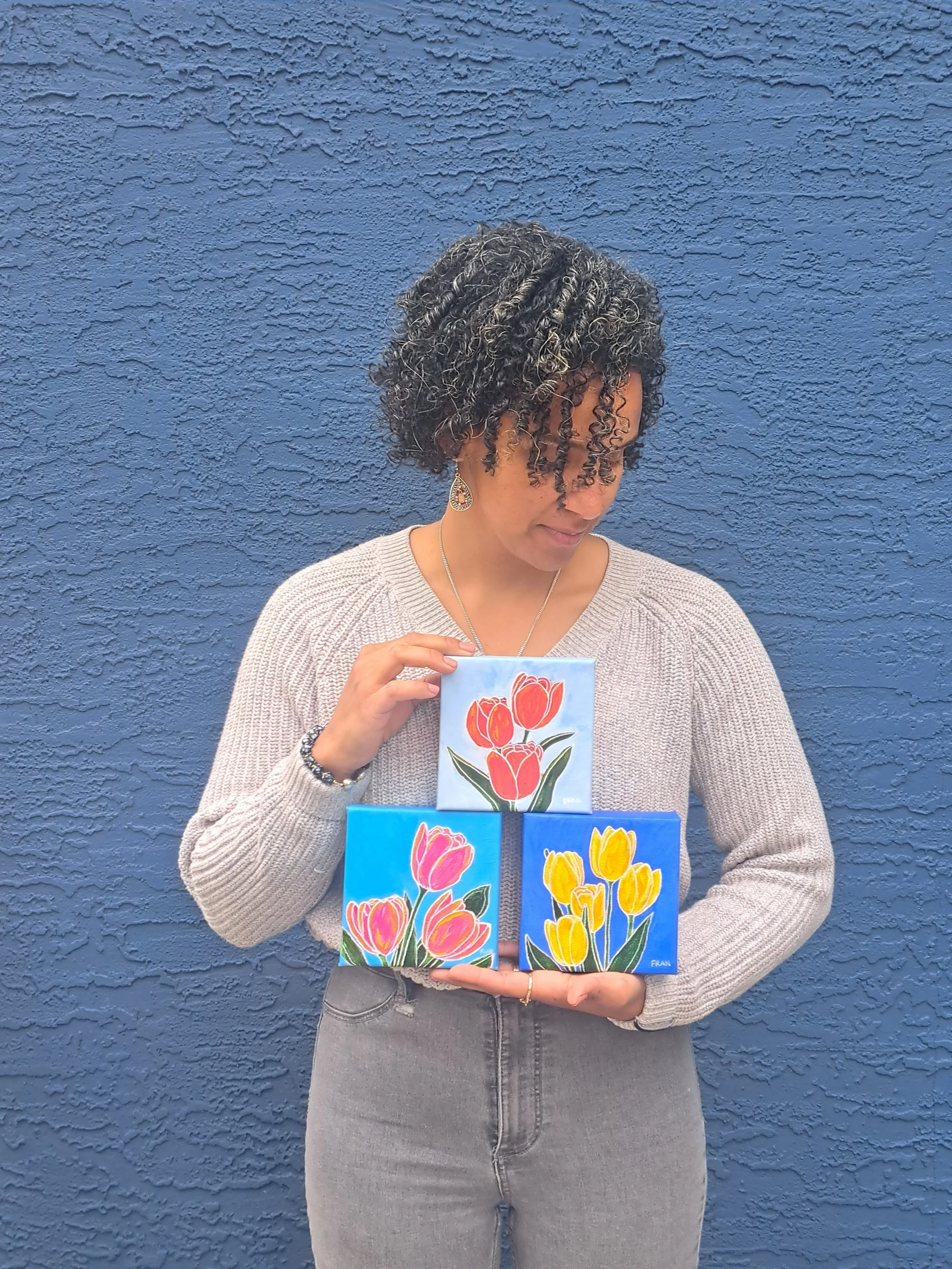 A woman with curly hair wearing a beige sweater and gray jeans holding three framed paintings of colorful tulips against a blue textured wall.