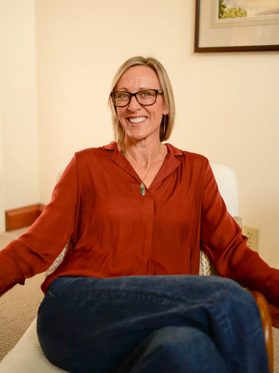 A woman with blonde hair, glasses, and a red blouse sitting on a beige armchair, smiling at the camera in a room with framed art on the wall.