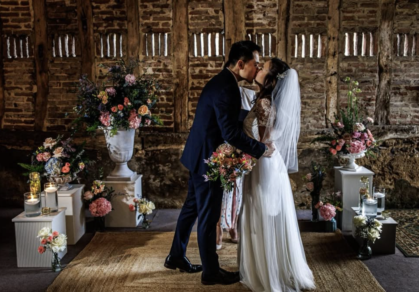 A bride and groom share a kiss during their wedding ceremony in a rustic setting with exposed brick and wooden beams. The bride wears a white lace gown and veil, and the groom is in a dark suit. Floral arrangements and candles decorate the space.