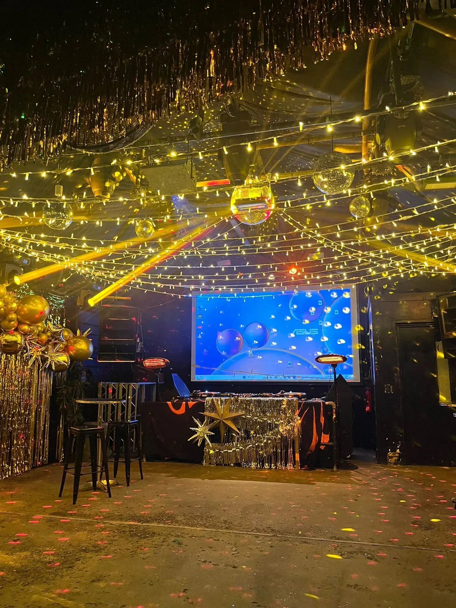 Dance floor decorated with gold balloons, hanging gold tinsel, and string lights, with a DJ booth in the background and a large screen displaying a blue and white display, indicating a party or celebration in a nightclub or event space.