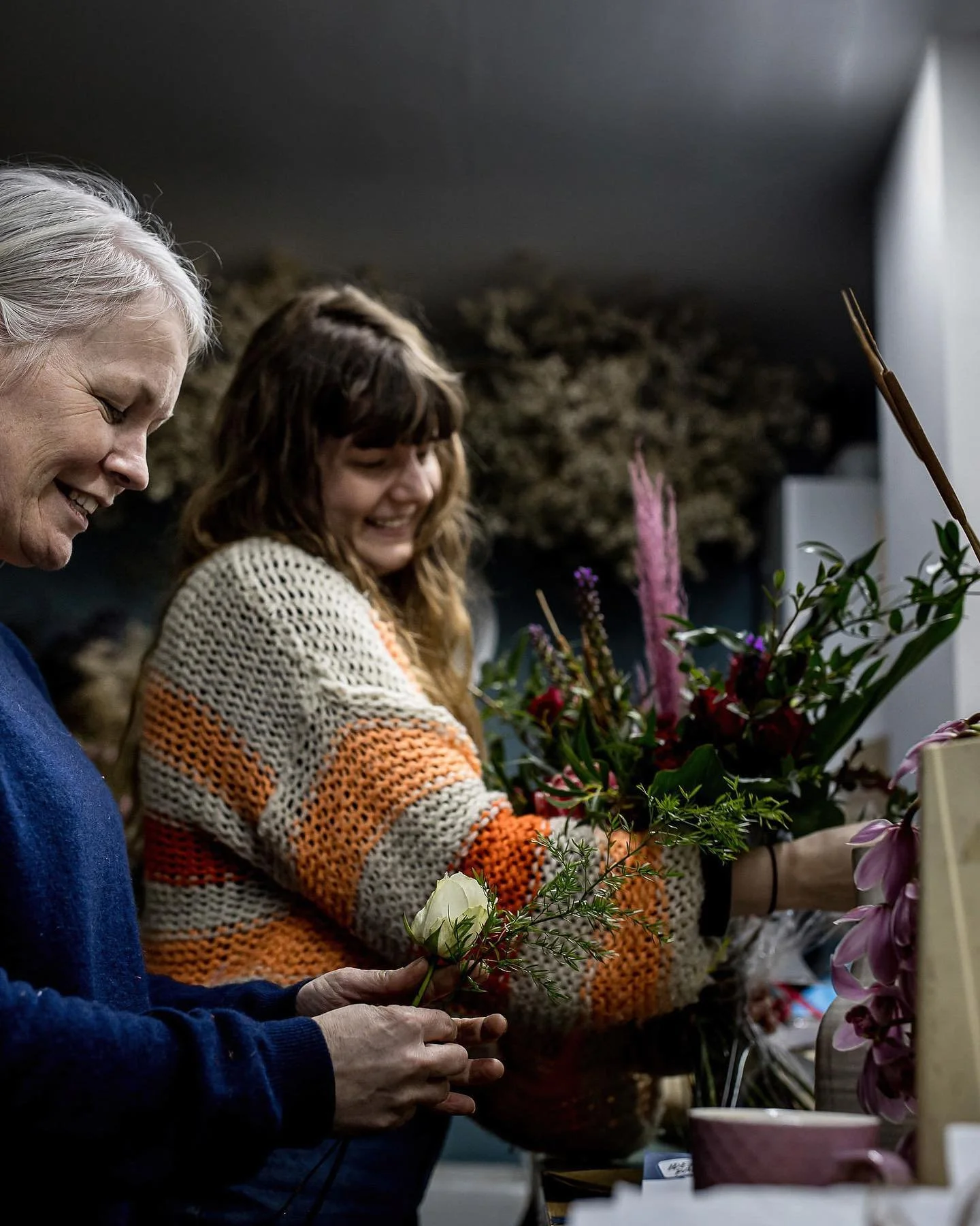 Two women smiling and working together on arranging flowers. One older woman with gray hair is holding a white flower, and a younger woman with brown, curly hair is arranging a large bouquet of colorful flowers.