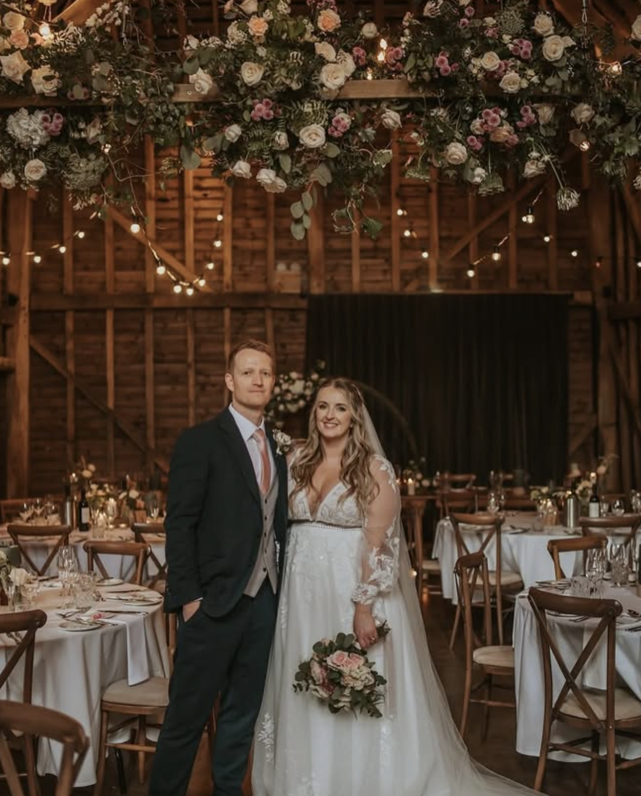 A bride and groom standing together inside a rustic wedding venue decorated with hanging flowers and string lights. The bride is holding a bouquet of flowers, and the groom is dressed in a suit and tie.