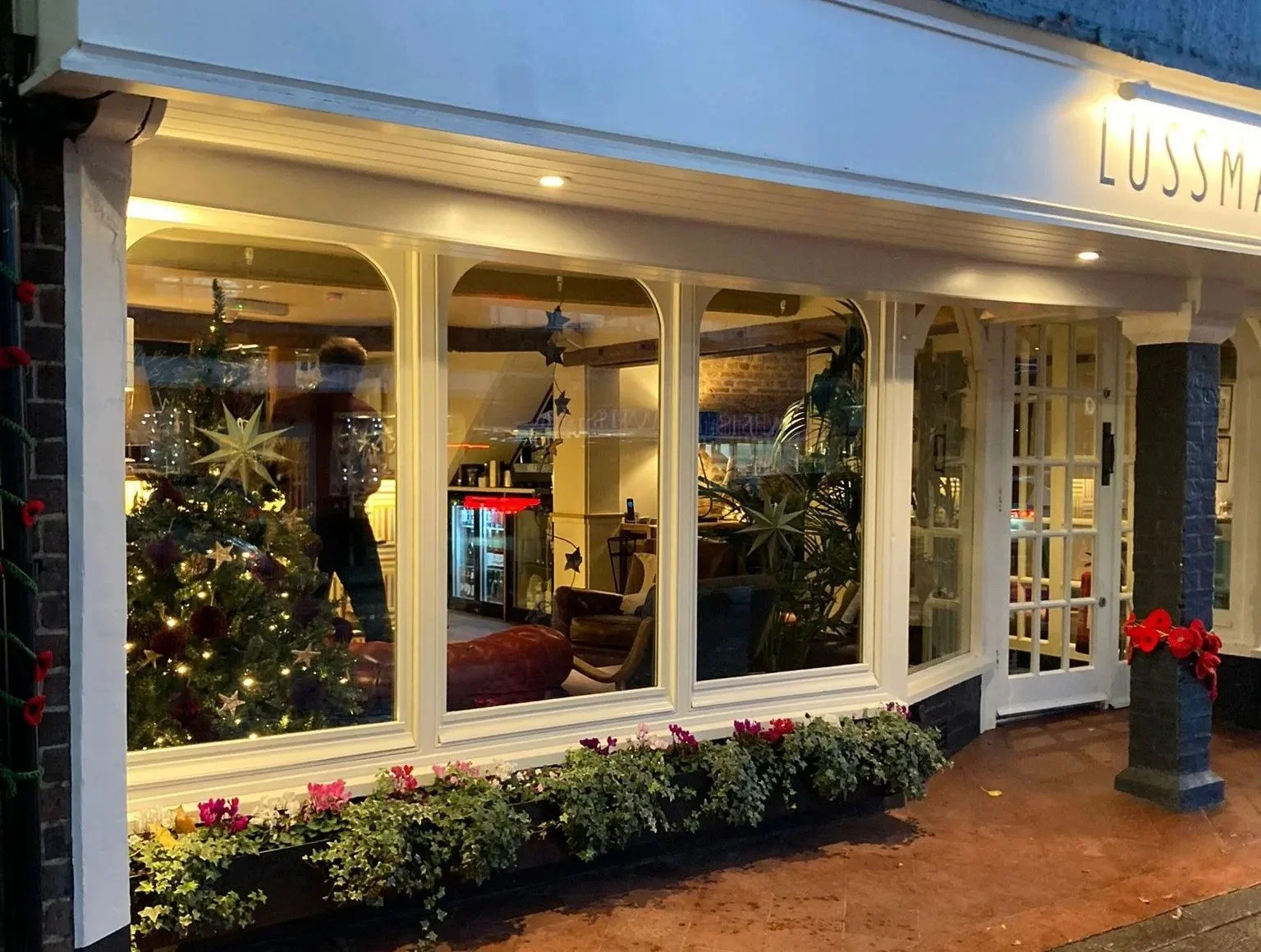 View of a cozy decorated restaurant window with a Christmas tree inside, fairy lights, and flowers outside, during evening hours.