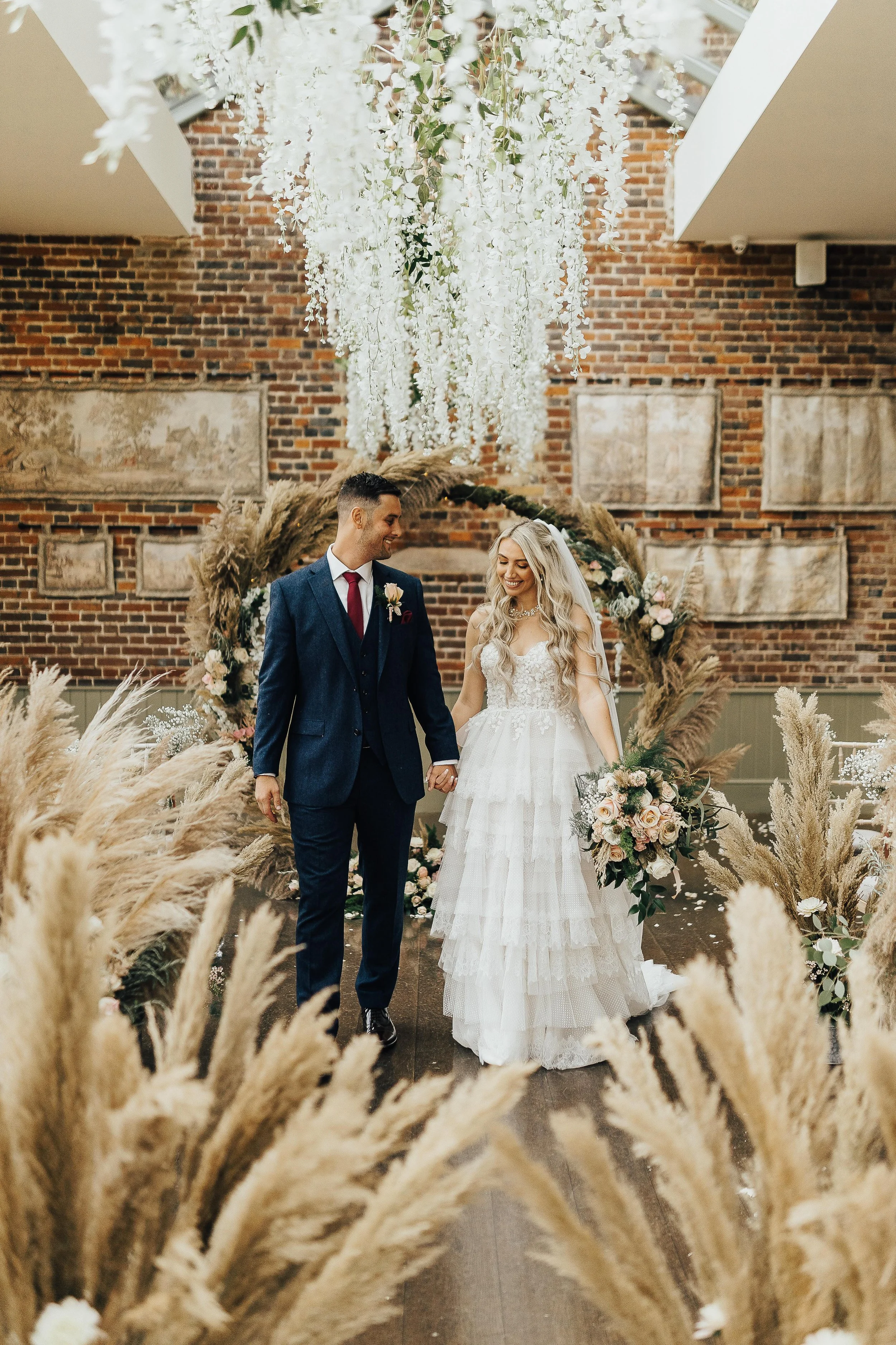 A bride and groom walking hand in hand through a decorated venue with pampas grass, floral arrangements, and a brick wall in the background.