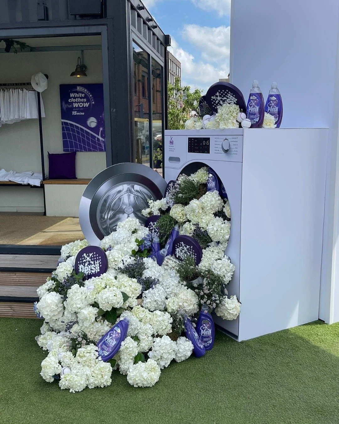 A washing machine filled with white flowers and bottles of Persil laundry detergent outside a building with a seating area and a sign about cleaning clothes.