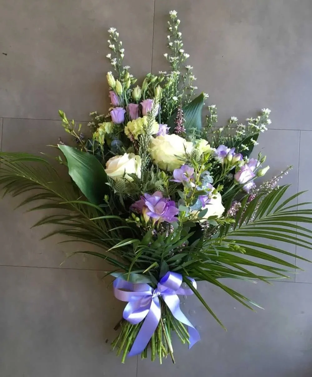 A funeral flower arrangement with white, purple, and light green flowers, accented with green leaves, tied with a purple ribbon.