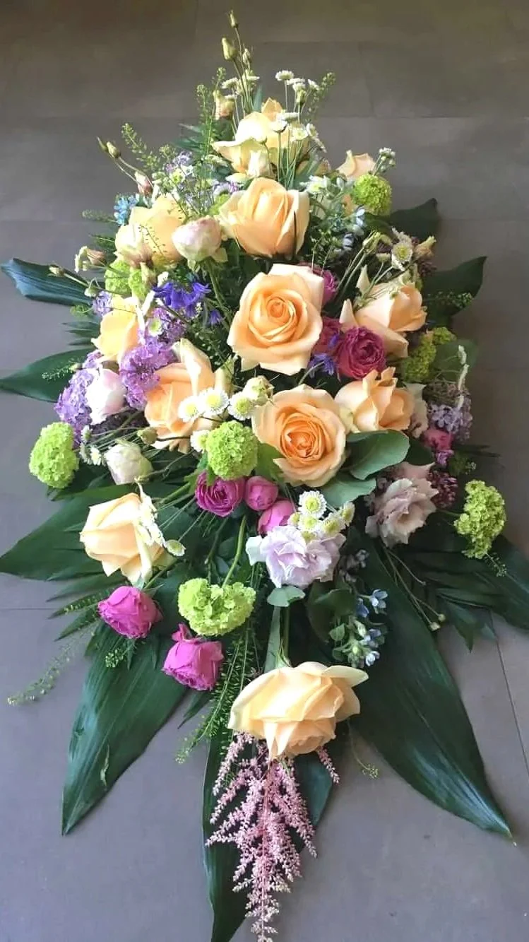 Colorful floral arrangement with peach roses, purple and pink flowers, white daisies, and green foliage on a dark surface.