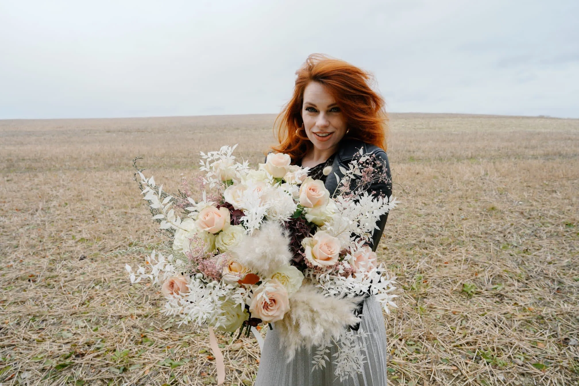Woman with red hair holding a large bouquet of white and pale pink flowers in an open field under a cloudy sky.