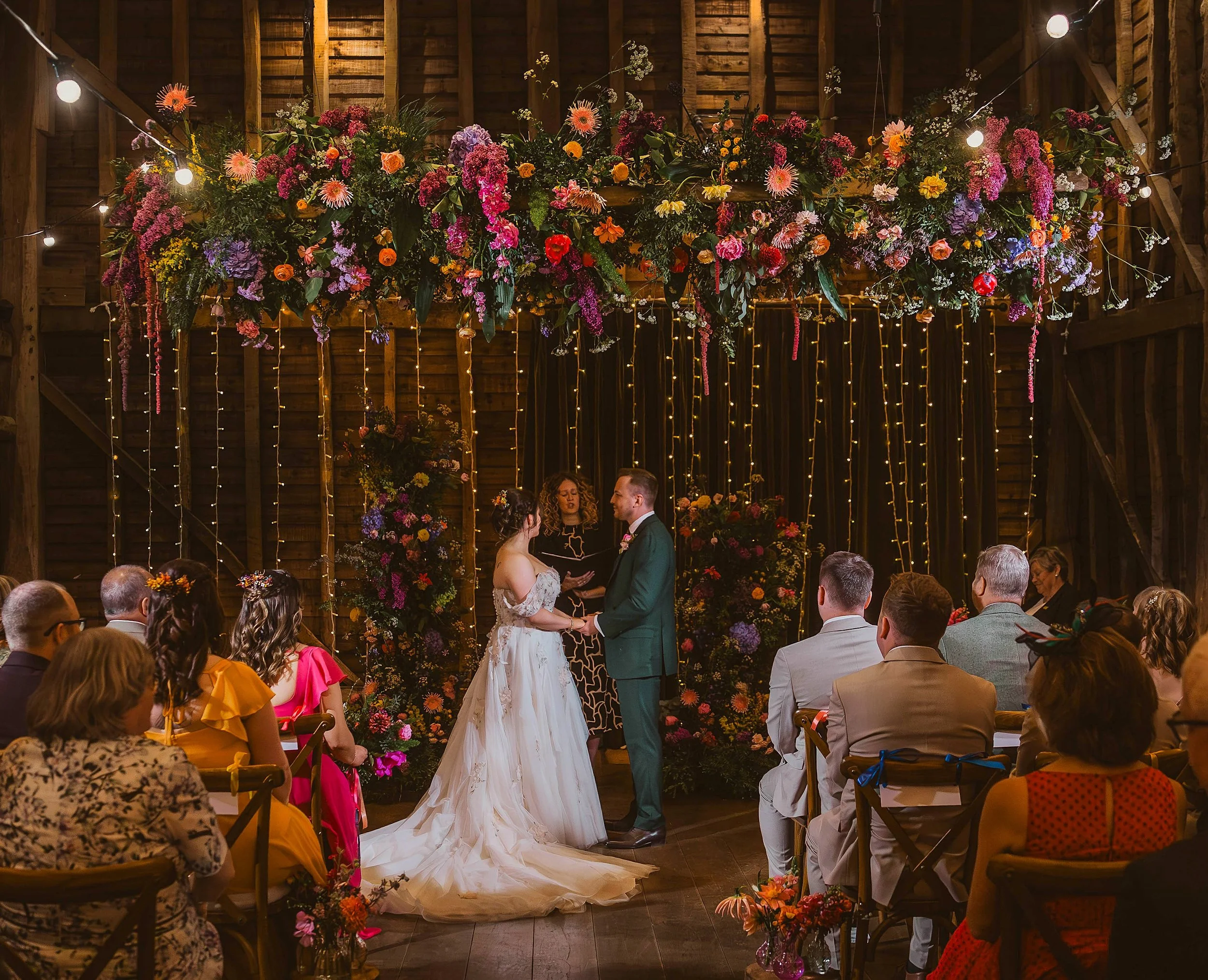 A wedding ceremony with a couple standing in front of an officiant, holding hands, in a rustic barn with floral decorations and string lights.