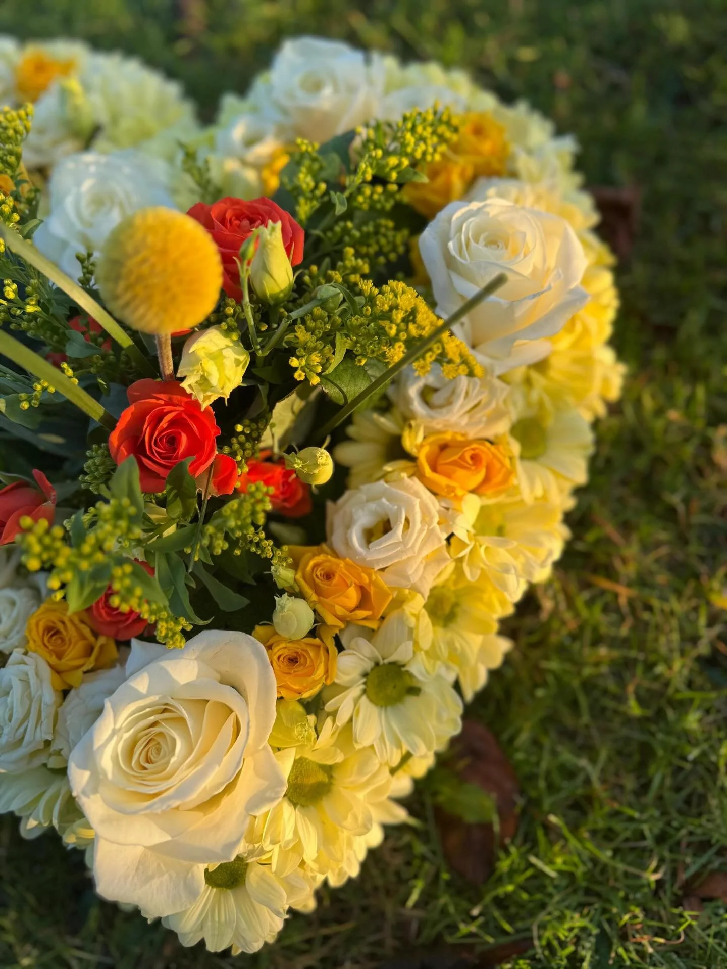 A colorful funeral flower arrangement on grass including white roses, yellow roses, red roses, and yellow filler flowers.