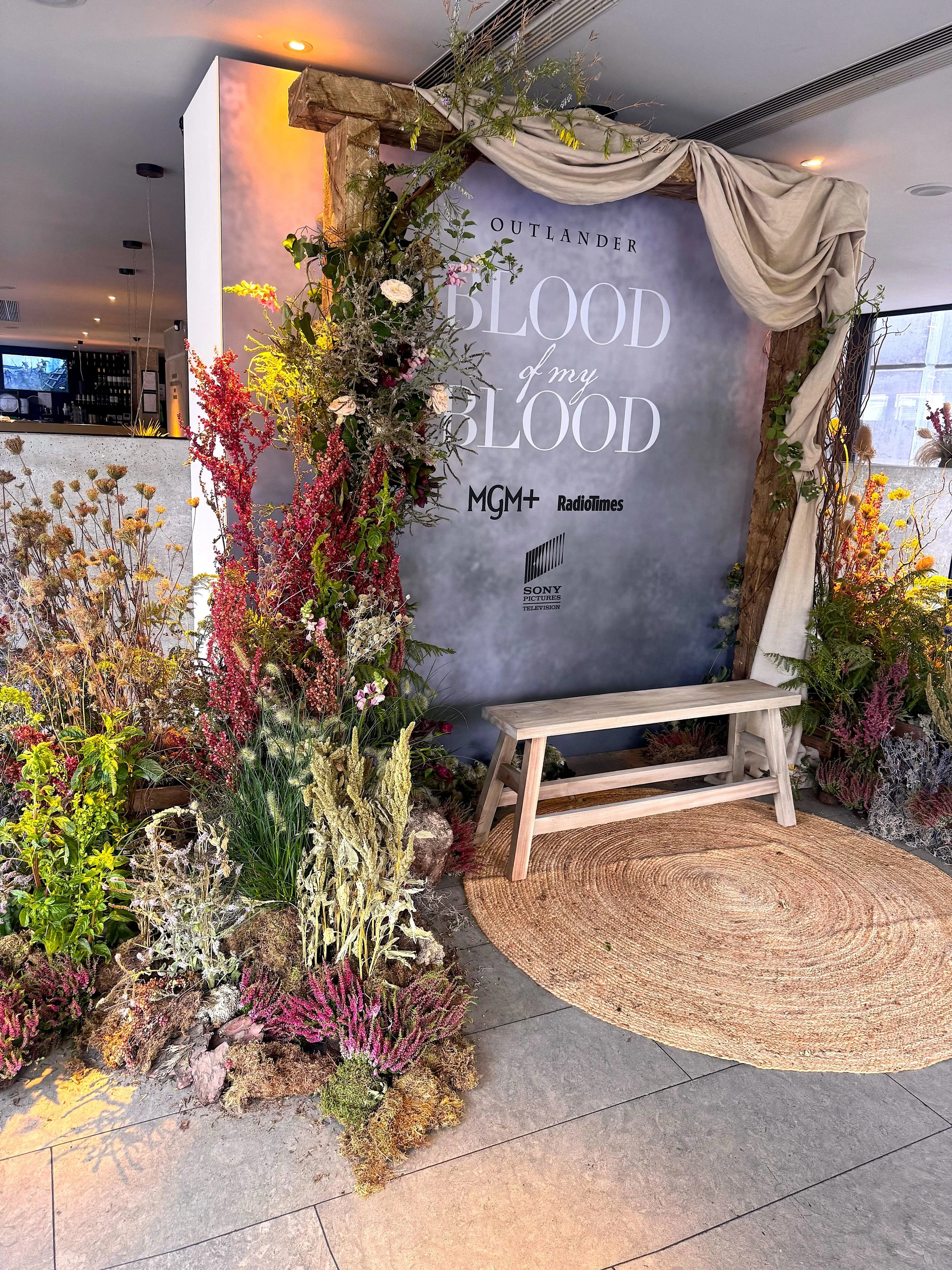 Decorative floral display with a wooden bench, a woven rug, and a backdrop with the text "Outlander Blood of my Blood" along with logos for MGM+, RadioTimes, and Sony Pictures Television.