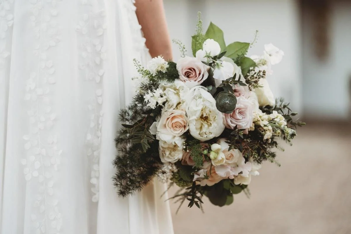 A bride holding a bouquet of white and blush pink roses, peonies, and greenery at a wedding.