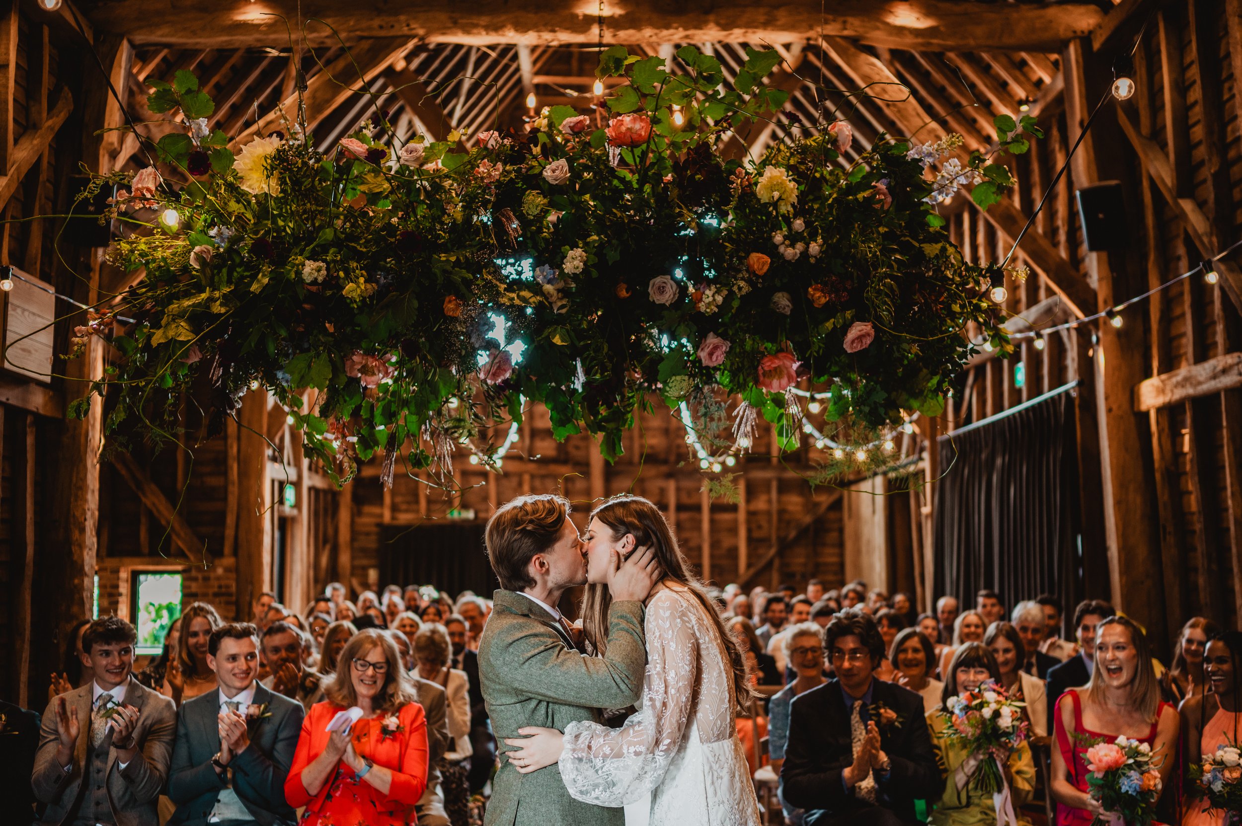 A wedding ceremony takes place in a rustic barn with wooden beams. The bride and groom kiss under an elaborate floral arrangement hanging from the ceiling, surrounded by seated guests smiling and clapping.