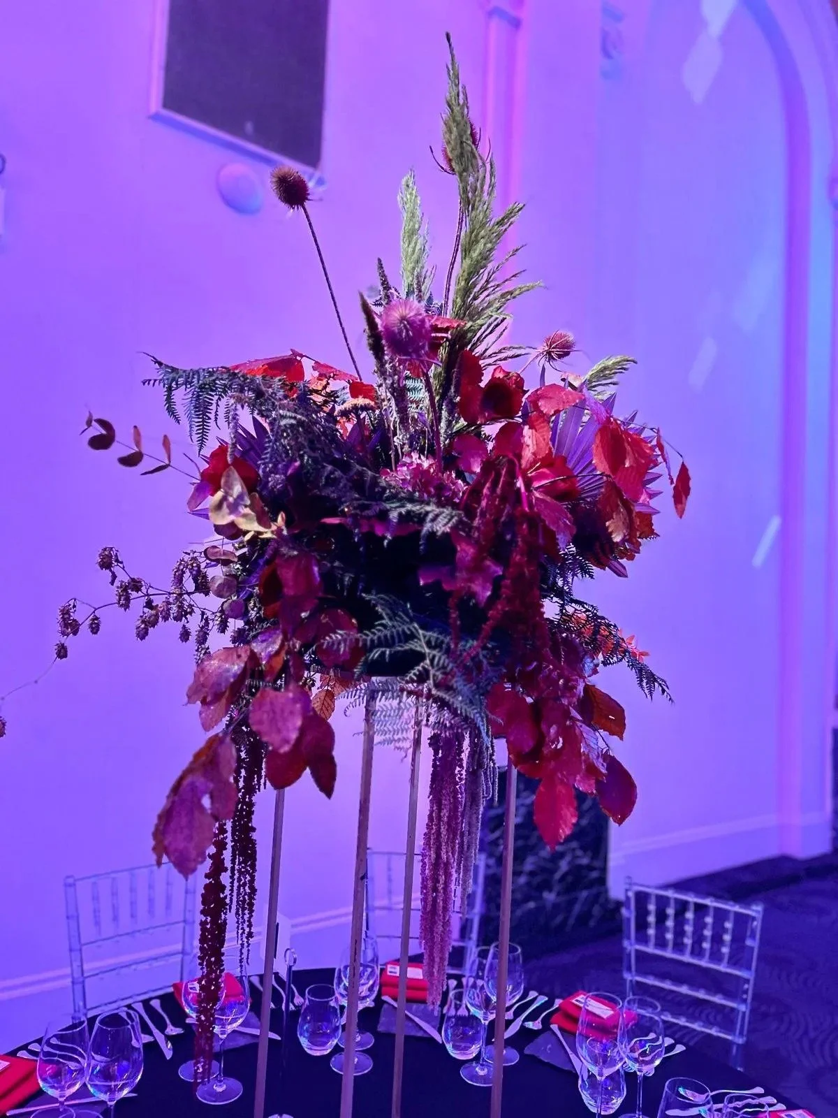 Tall floral centerpiece with red, purple, and green foliage on a banquet table with wine glasses and red napkins, set against a purple-lit background.