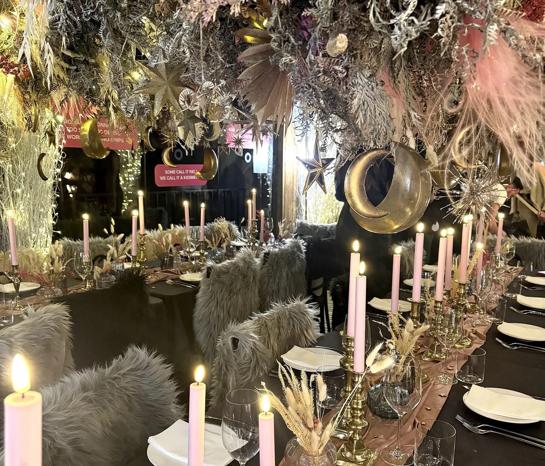 A festive dining setup with pink candles, fluffy faux fur chairs, and hanging gold and silver decorations including crescent moons, stars, and sparkles amidst pink and white floral arrangements.