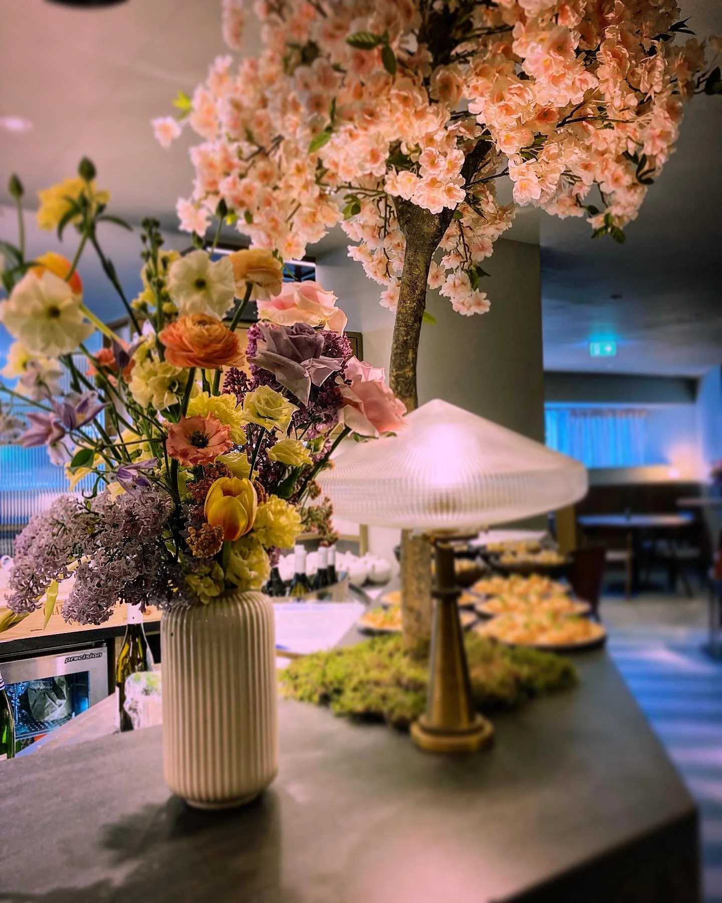 A floral arrangement with pink cherry blossoms in a vase, along with various colorful flowers, is placed next to a table lamp on a countertop.
