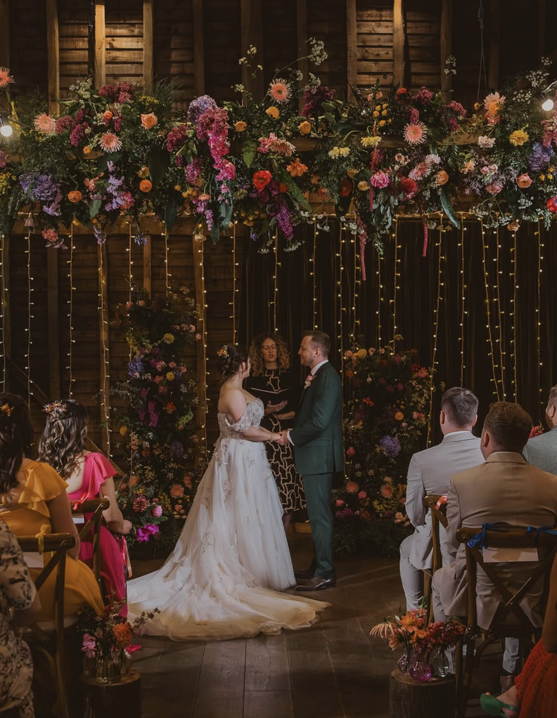A wedding ceremony taking place indoors with a bride and groom holding hands, surrounded by guests. The backdrop is decorated with an abundance of colorful flowers and string lights.